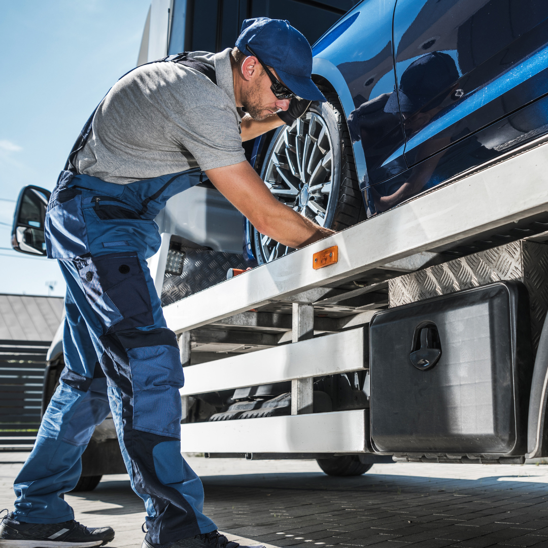 A man wearing a blue cap, sunglasses, and gray T-shirt working underneath a blue car on a flatbed truck, inspecting or repairing the vehicle.