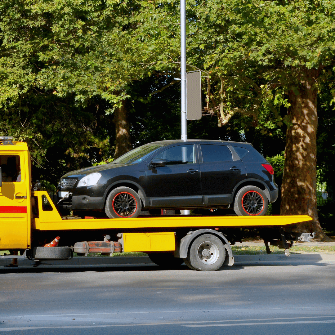 Black compact hatchback car on a yellow tow truck on a city street with green trees in the background.