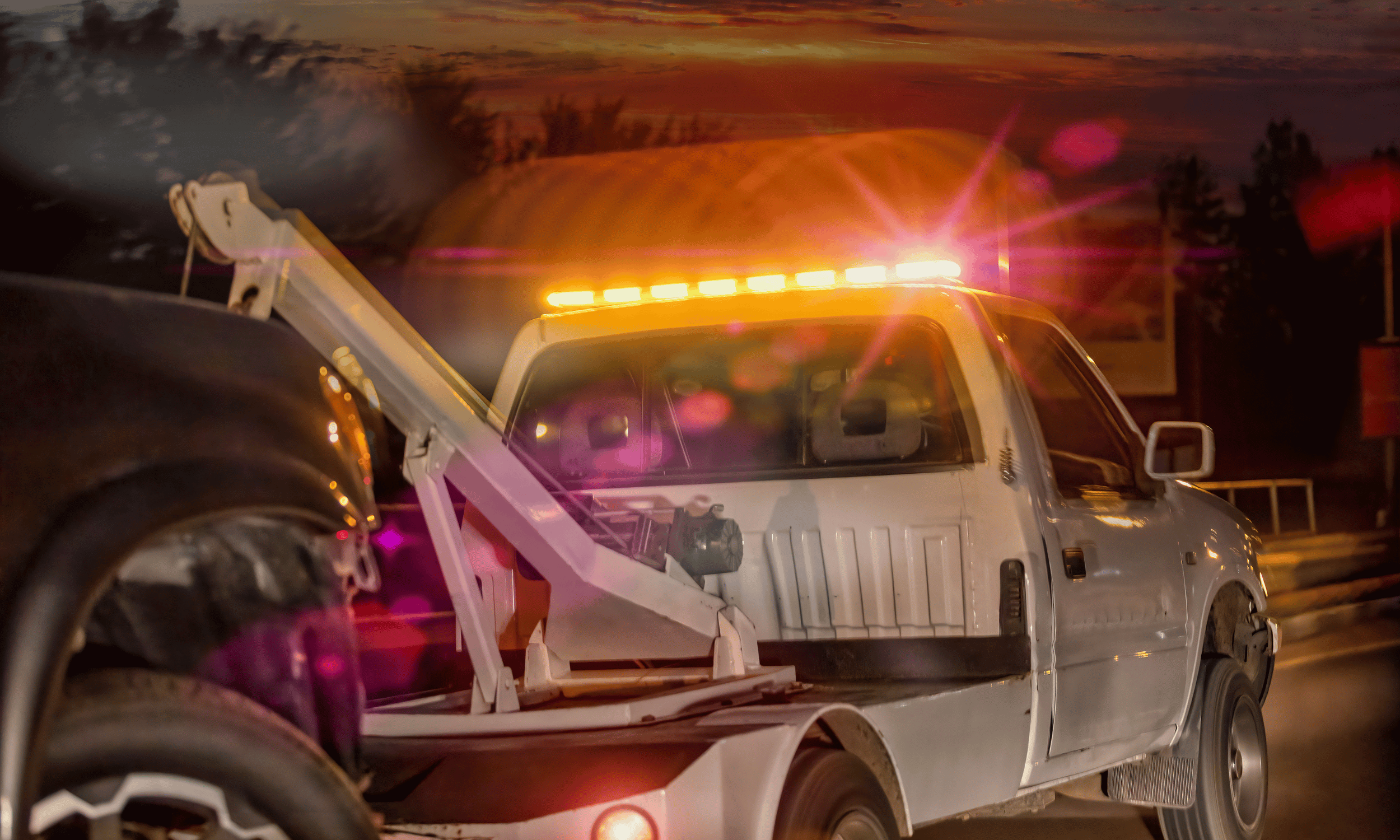 A tow truck at sunset with flashing yellow warning lights, preparing to tow a black vehicle.