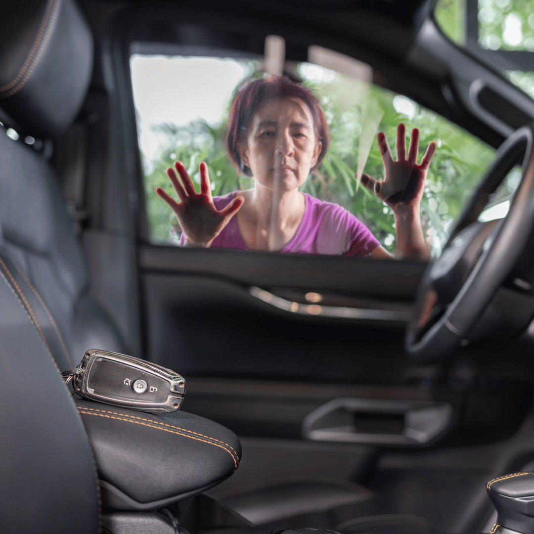 A woman with short dark hair, wearing a pink shirt, is standing outside a car, pressing her hands against a car window, with trees and greenery visible in the background.