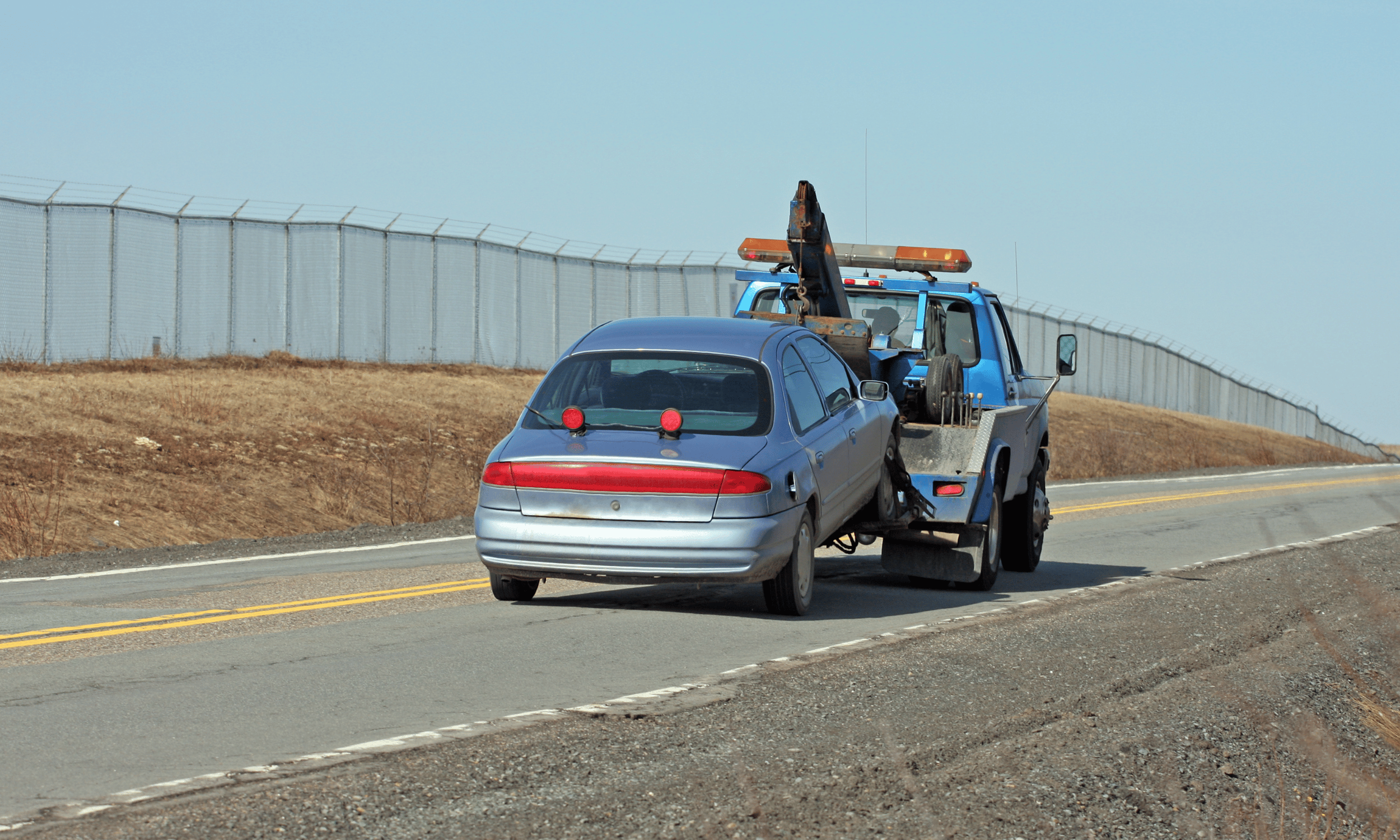 Tow truck towing a damaged silver car on a rural road with a metal fence along the roadside.