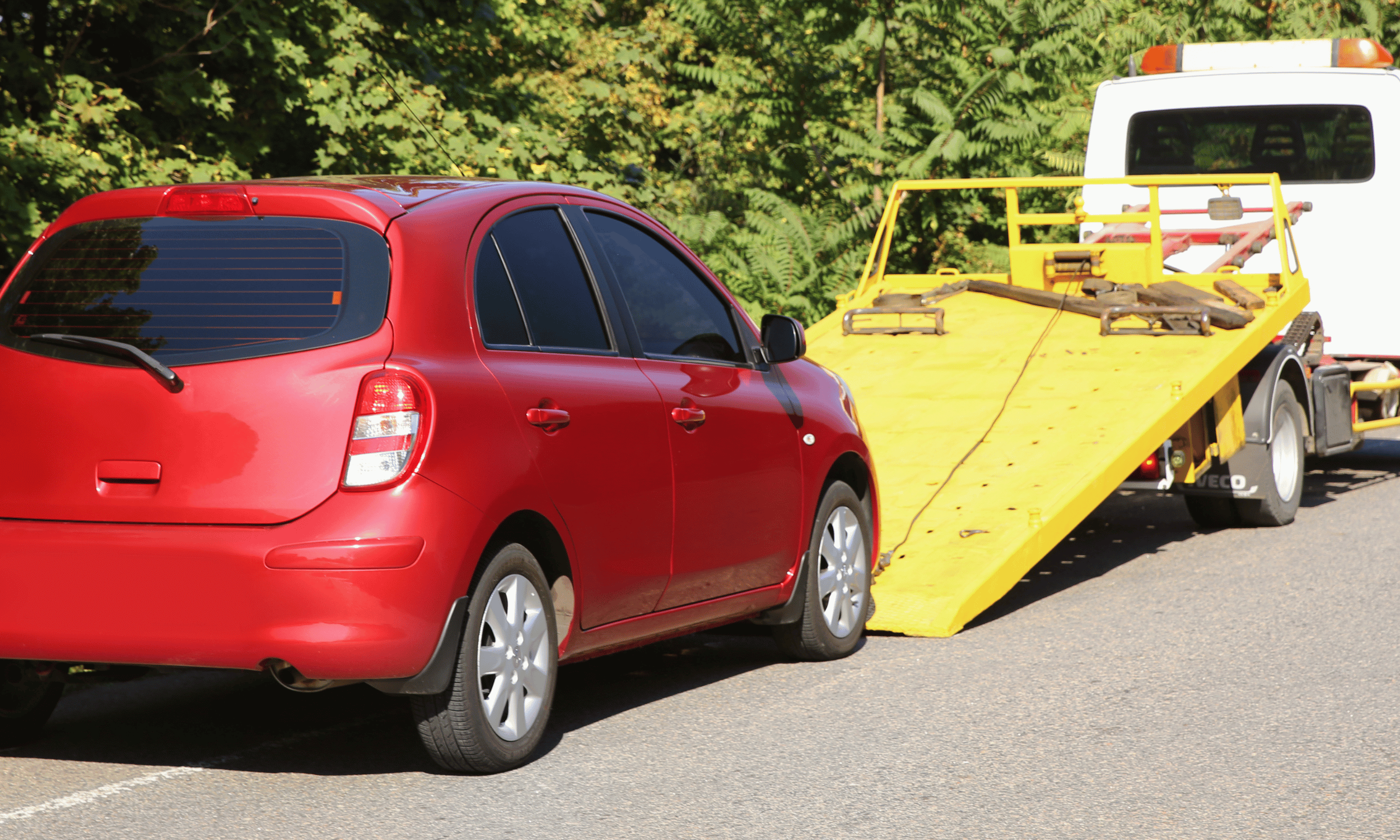 A red car is being loaded onto a yellow tow truck on a paved road with green foliage in the background.
