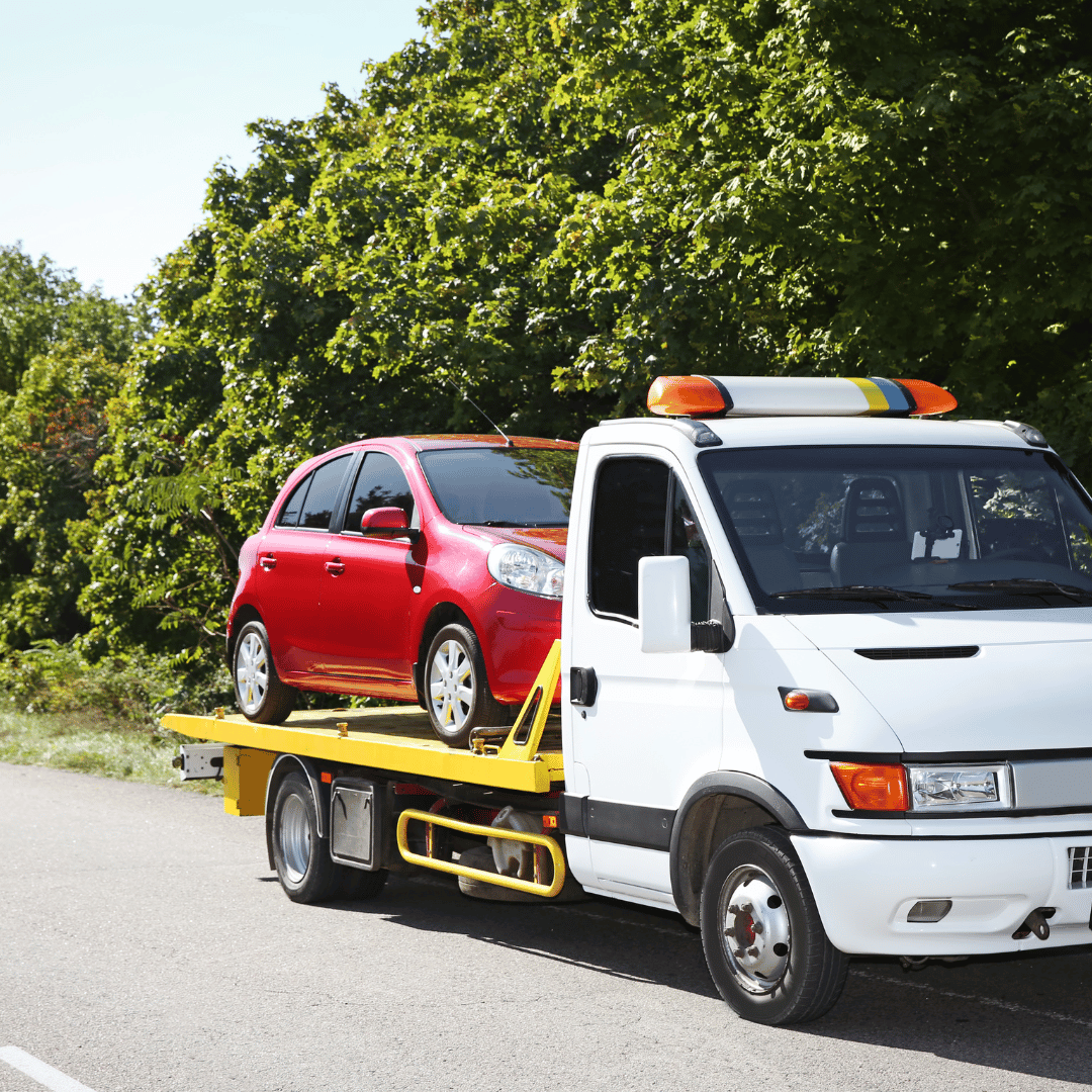 White tow truck transporting a red compact car on a yellow flatbed, parked on the side of a road with green trees in the background.