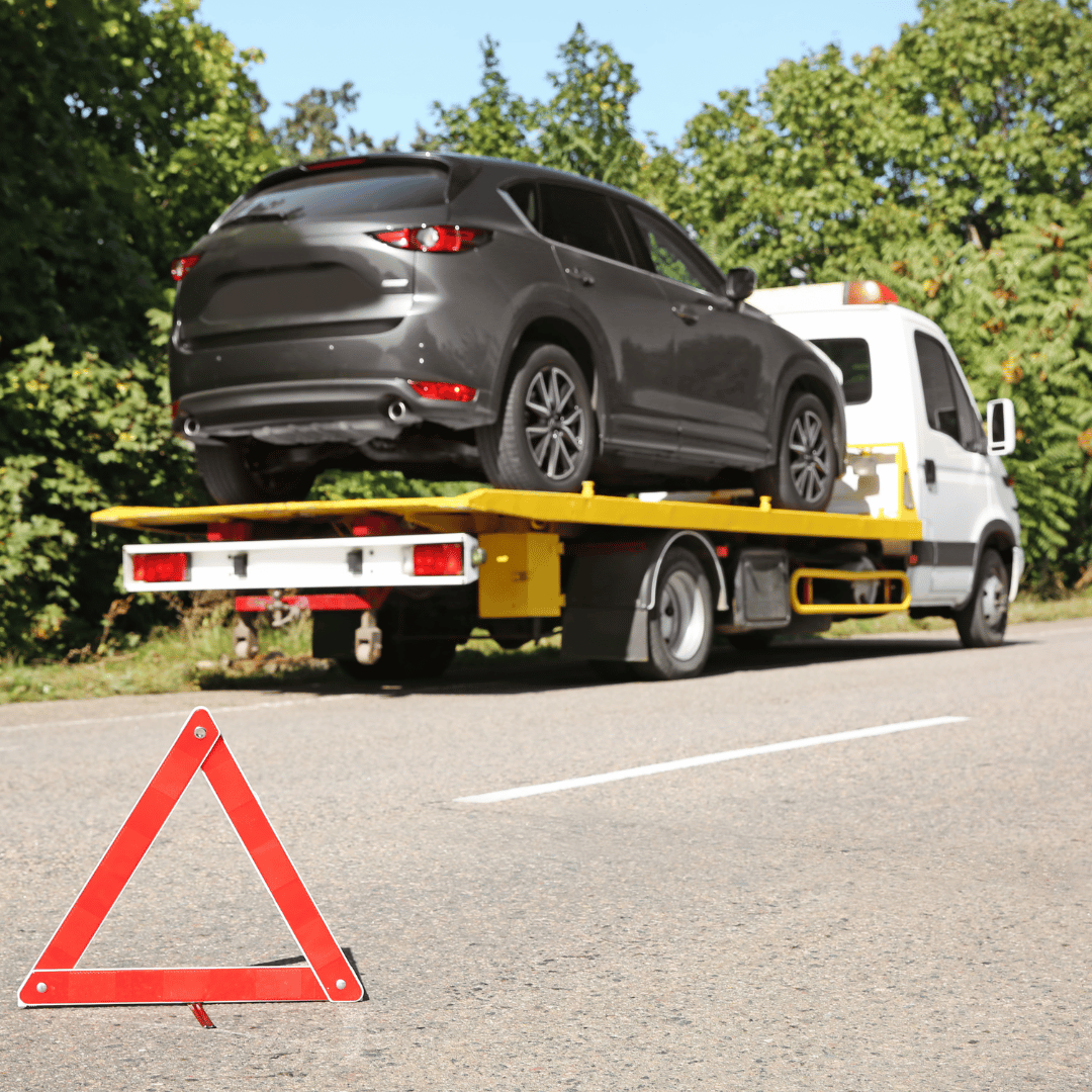 A flatbed tow truck carrying a black SUV on a road with trees in the background and a warning triangle placed on the ground in front.