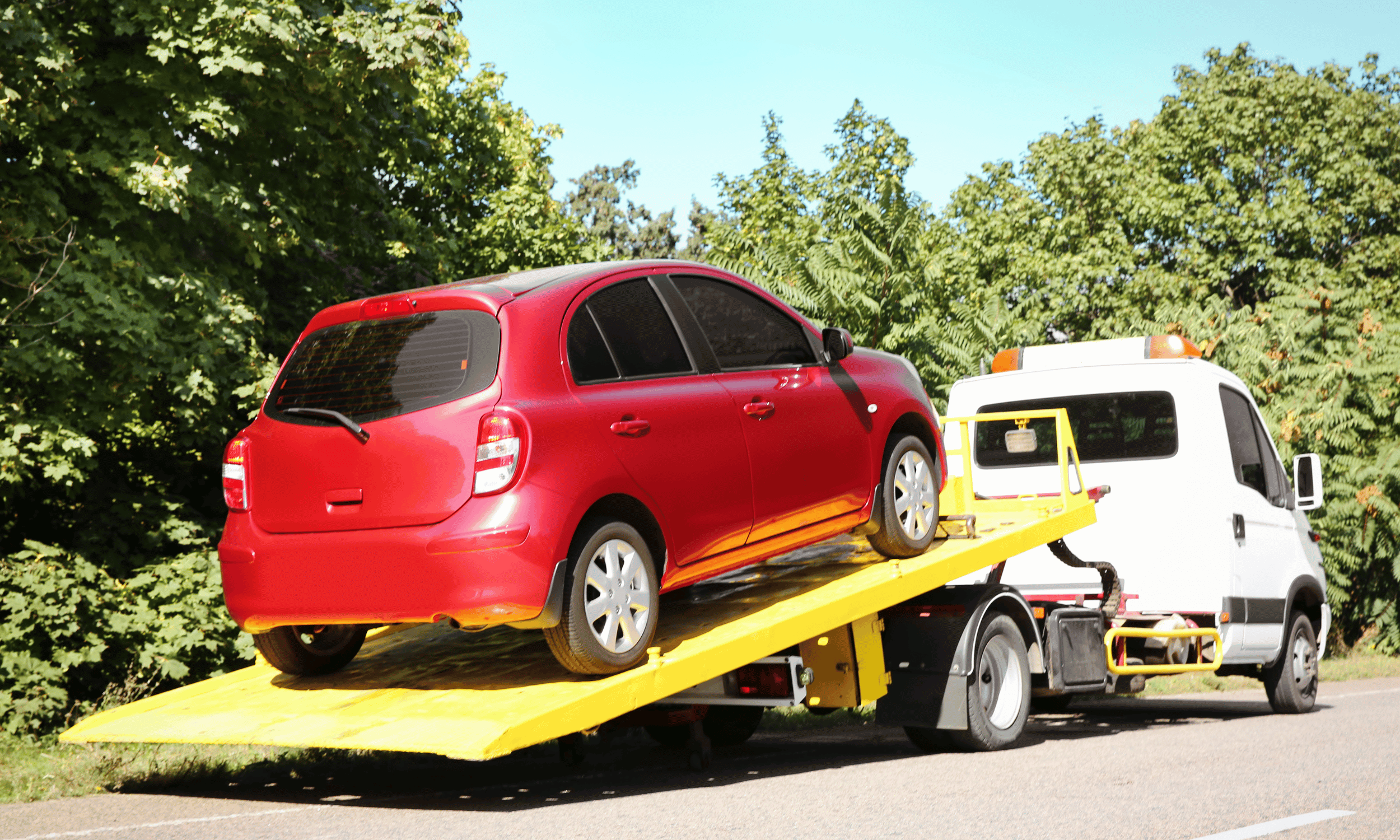 A red compact car being loaded onto a yellow tow truck on the side of a road with trees in the background.