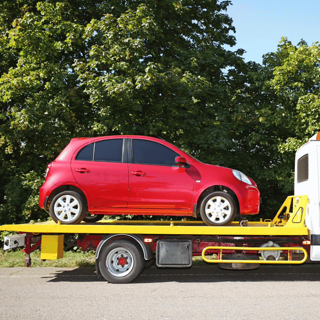 Red compact car being transported on a flatbed tow truck during daytime, with green trees and clear blue sky in the background.