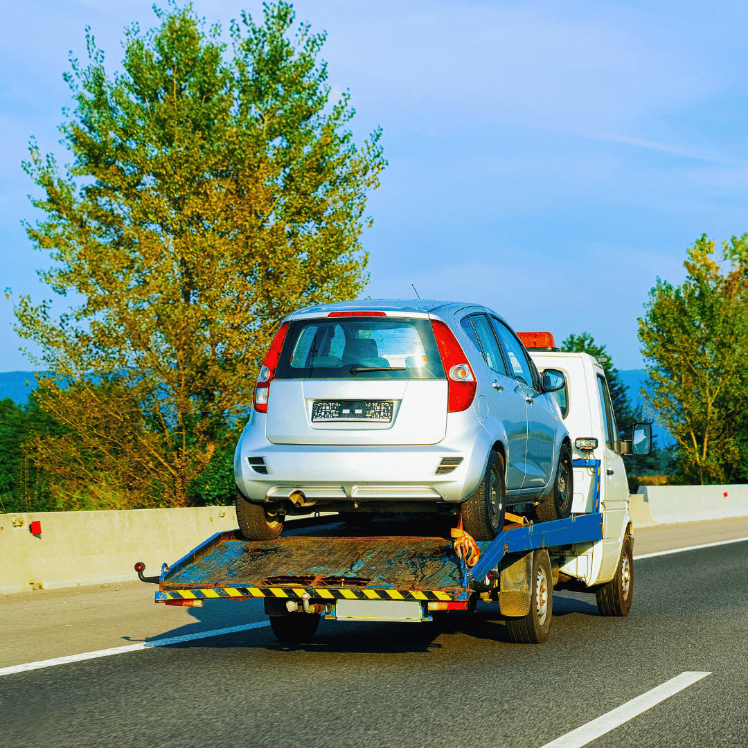 A flatbed tow truck transporting a silver hatchback car on a highway with trees and a blue sky in the background.