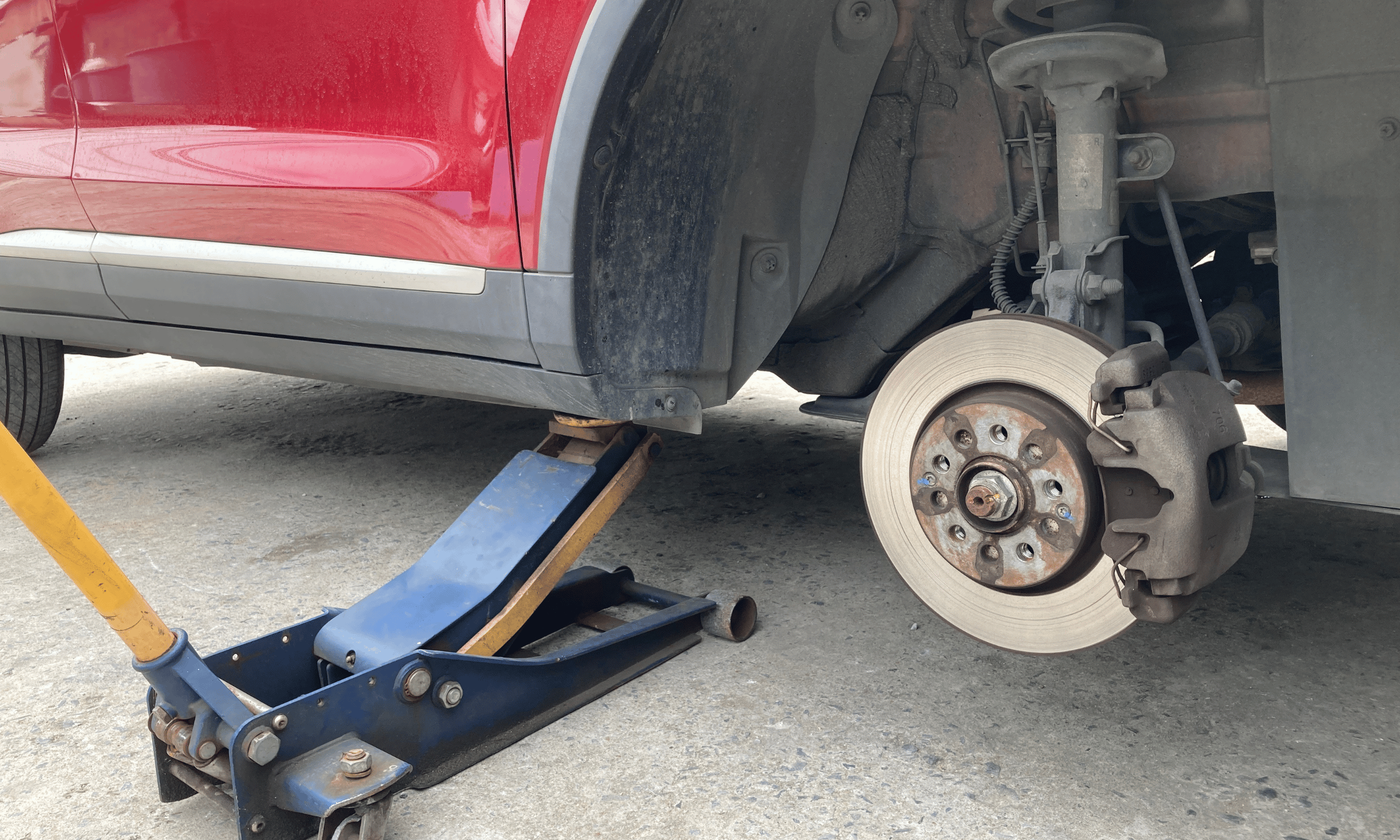 A red car is lifted with a hydraulic jack, with its front wheel removed exposing the brake caliper and rotor, on a paved surface.