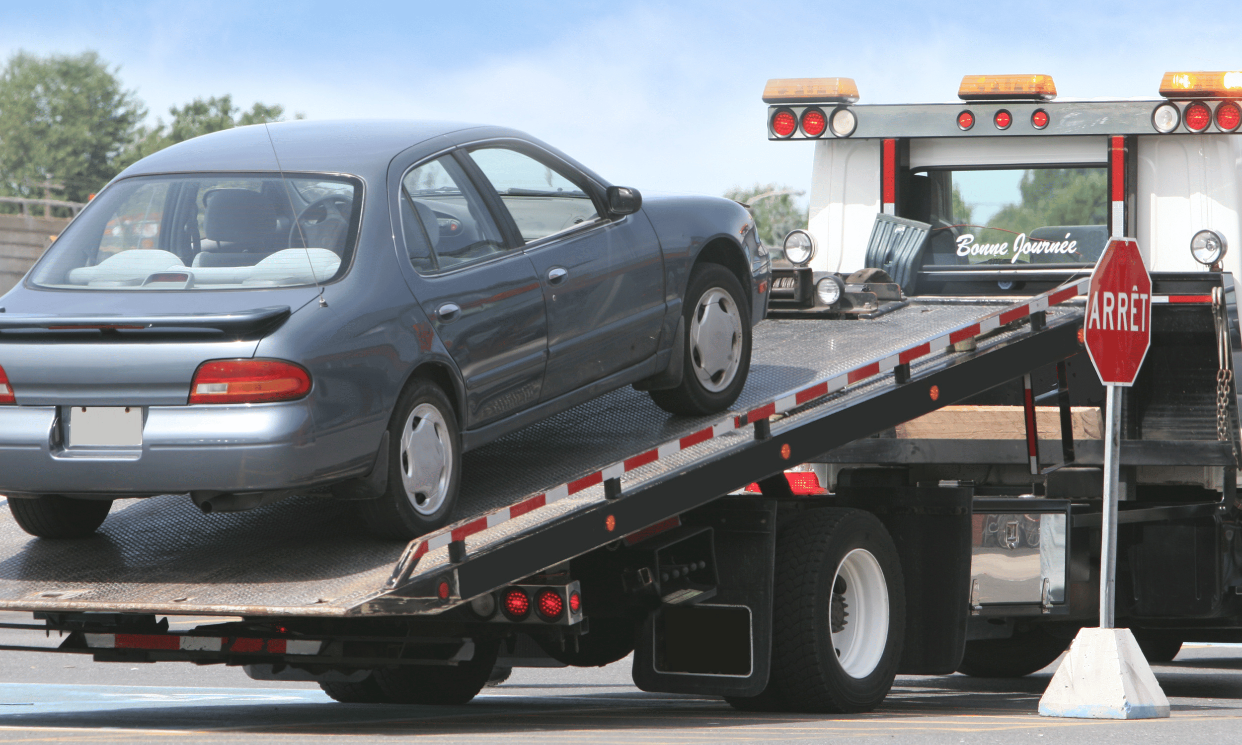 A silver car is on the back of a flatbed tow truck with a stop sign and a bilingual 'Good Day' message, 'Bonne Journée', displayed on the truck.