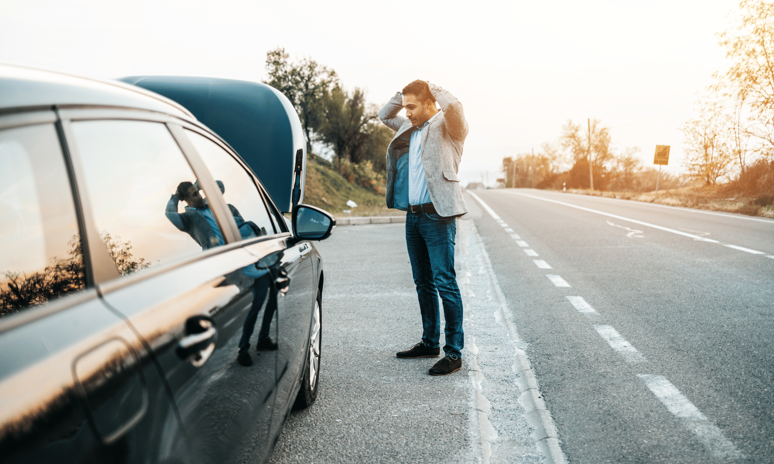 A man standing on the side of an empty road, inspecting his car with the hood open, during sunset.
