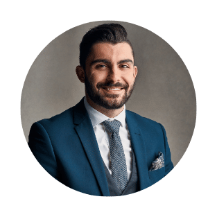 A man with dark hair and a beard wearing a blue suit, white shirt, and tie, smiling in front of a gray background.