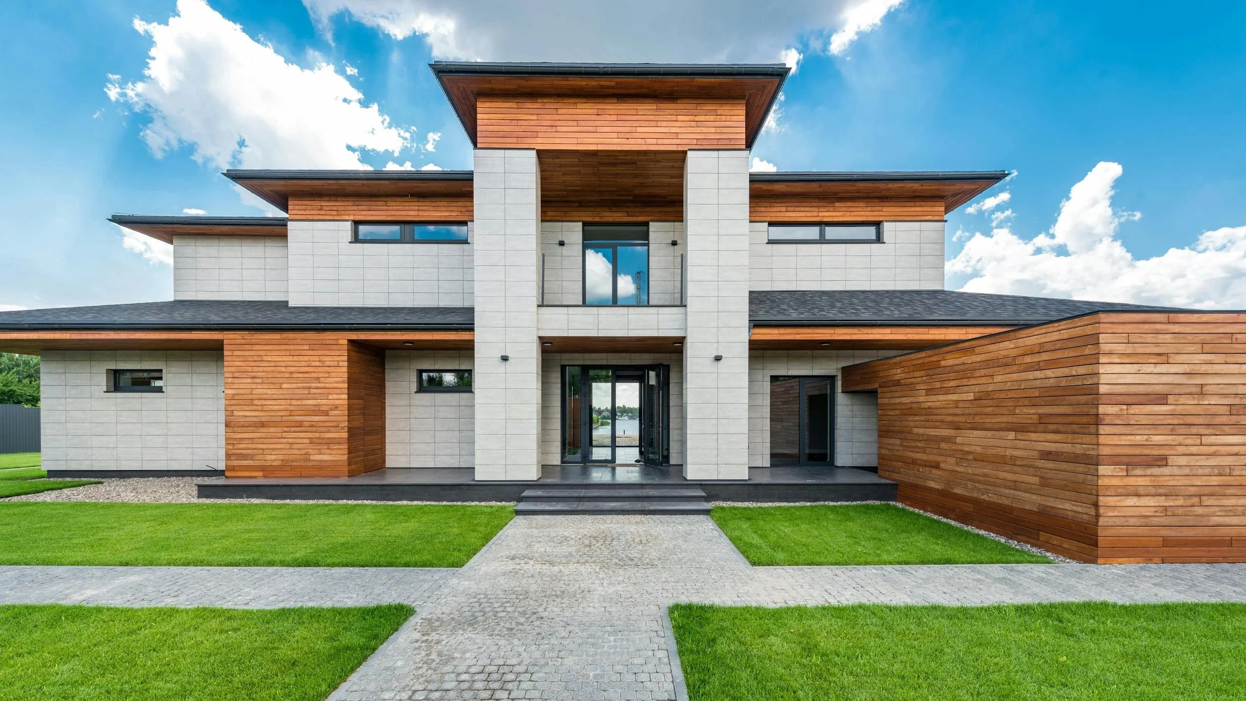 Modern house with wooden and light gray exterior, glass entry door, green lawn, cloudy sky.