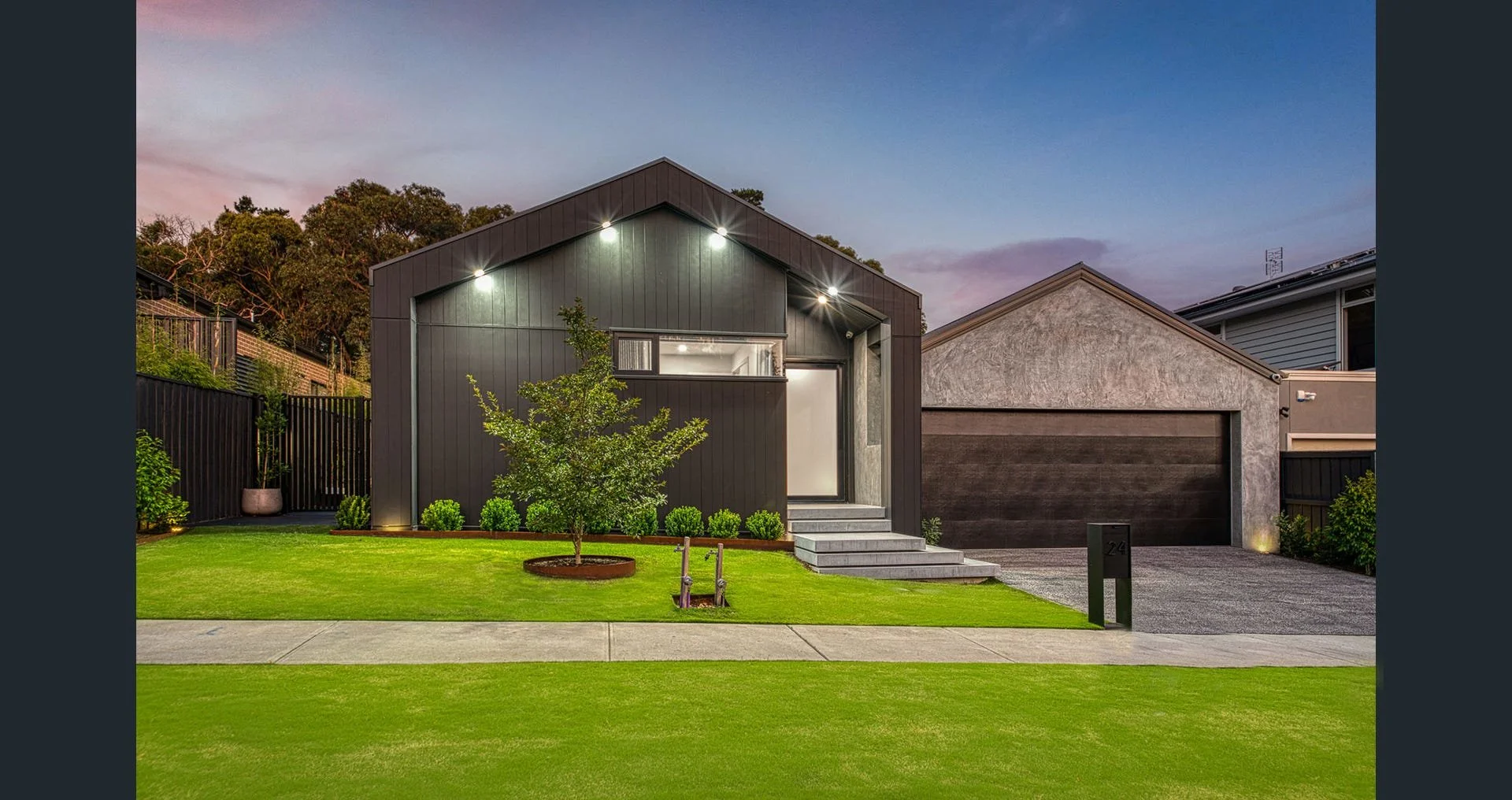 Modern house with dark exterior, well-lit front entrance, small front yard with green grass and a young tree, driveway, and neighboring houses, during dusk.