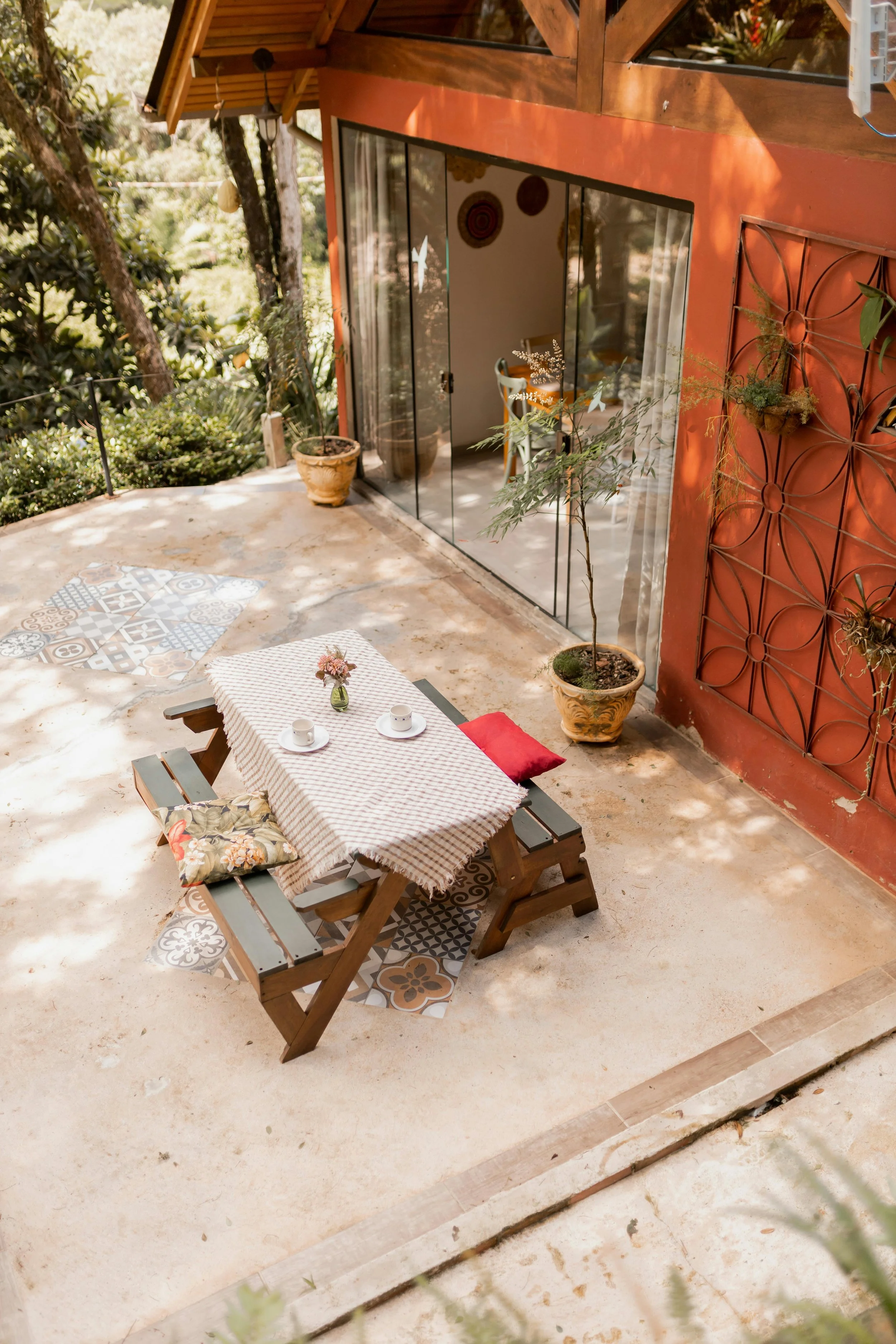 An outdoor patio area with a wooden table covered with a red and white checkered tablecloth, two cups, and a small vase with flowers. There are two benches with colorful cushions, potted plants, and decorative tiles on the floor. A glass sliding door leads inside, and there are trees and greenery surrounding the patio.