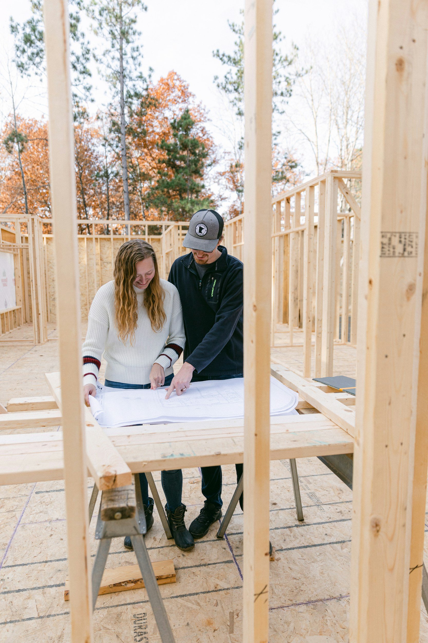 Two people, a young woman and a man, are standing over architectural blueprints on a table inside a new home under construction. The building's wooden framing is visible, and they are smiling while discussing the plans. 