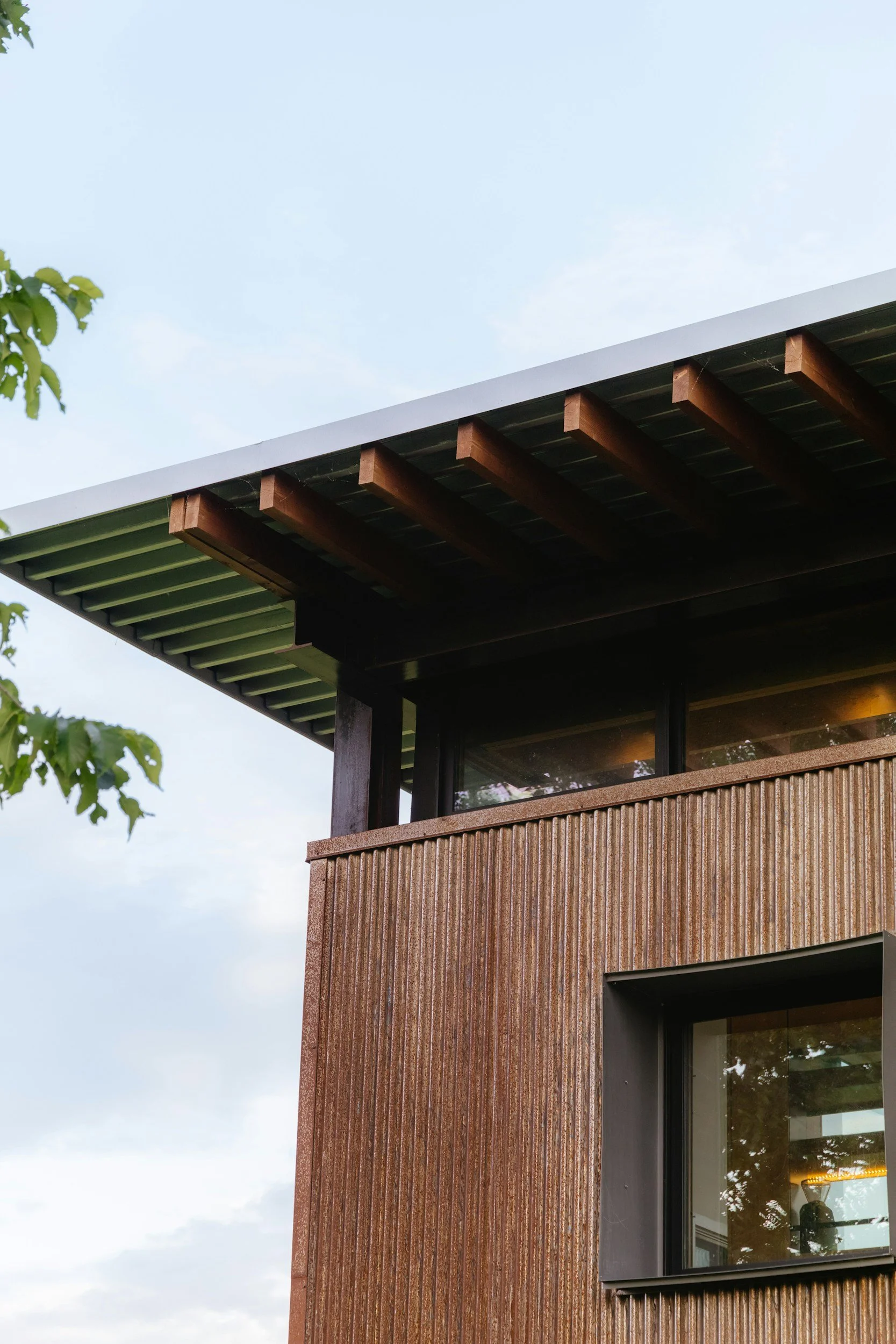 A modern building with a brown textured exterior wall, large glass window, and horizontal wooden beams supporting the eaves. Part of a tree with green leaves is visible on the left side. The sky is light blue with soft clouds.