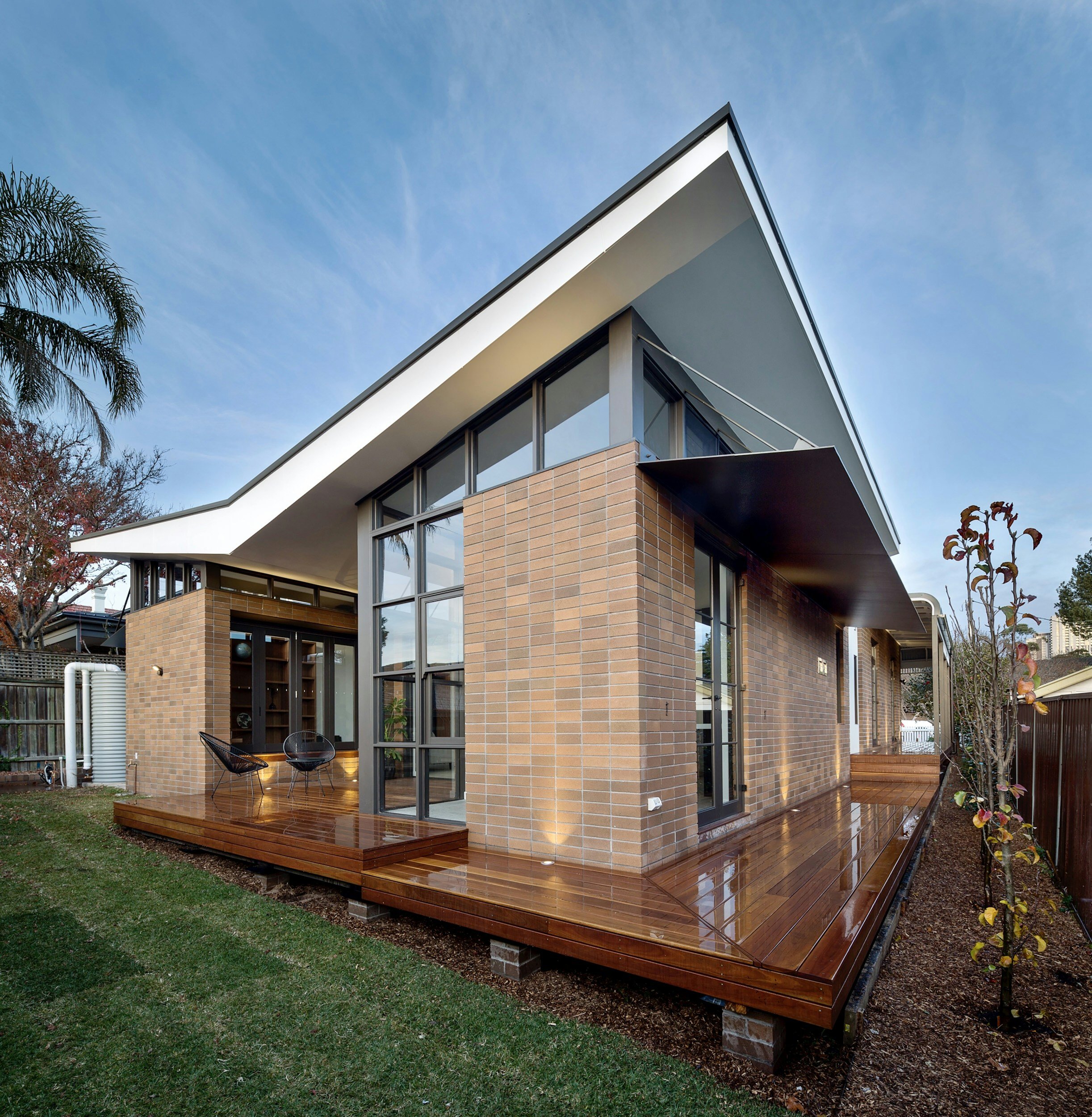 Modern house with a brick and glass exterior, a wooden deck, and a backyard with grass, trees, and a clear sky.