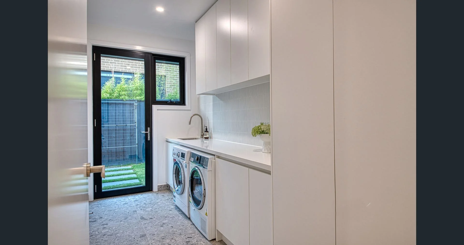 Modern laundry room with white cabinets, front-loading washer and dryer, a sink with a faucet, a countertop, a small potted plant, a window, and a glass door leading outside to a fenced yard with grass.
