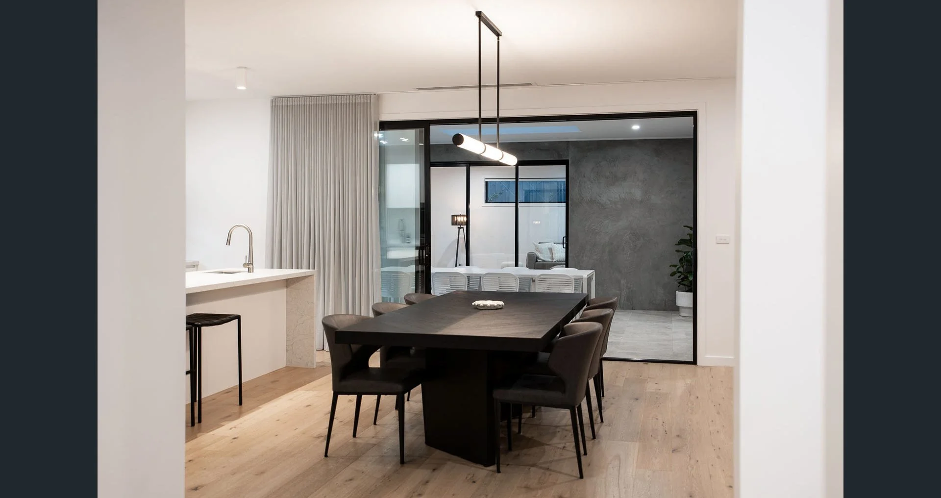 Modern dining area with a black table, six chairs, a pendant light, and a sliding glass door leading to a living space with a concrete wall and potted plant.