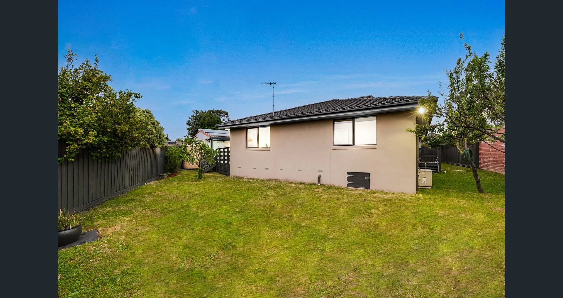Backyard with a grassy lawn, surrounded by a black fence, and a single-story house with beige walls and a dark roof, under a clear blue sky.