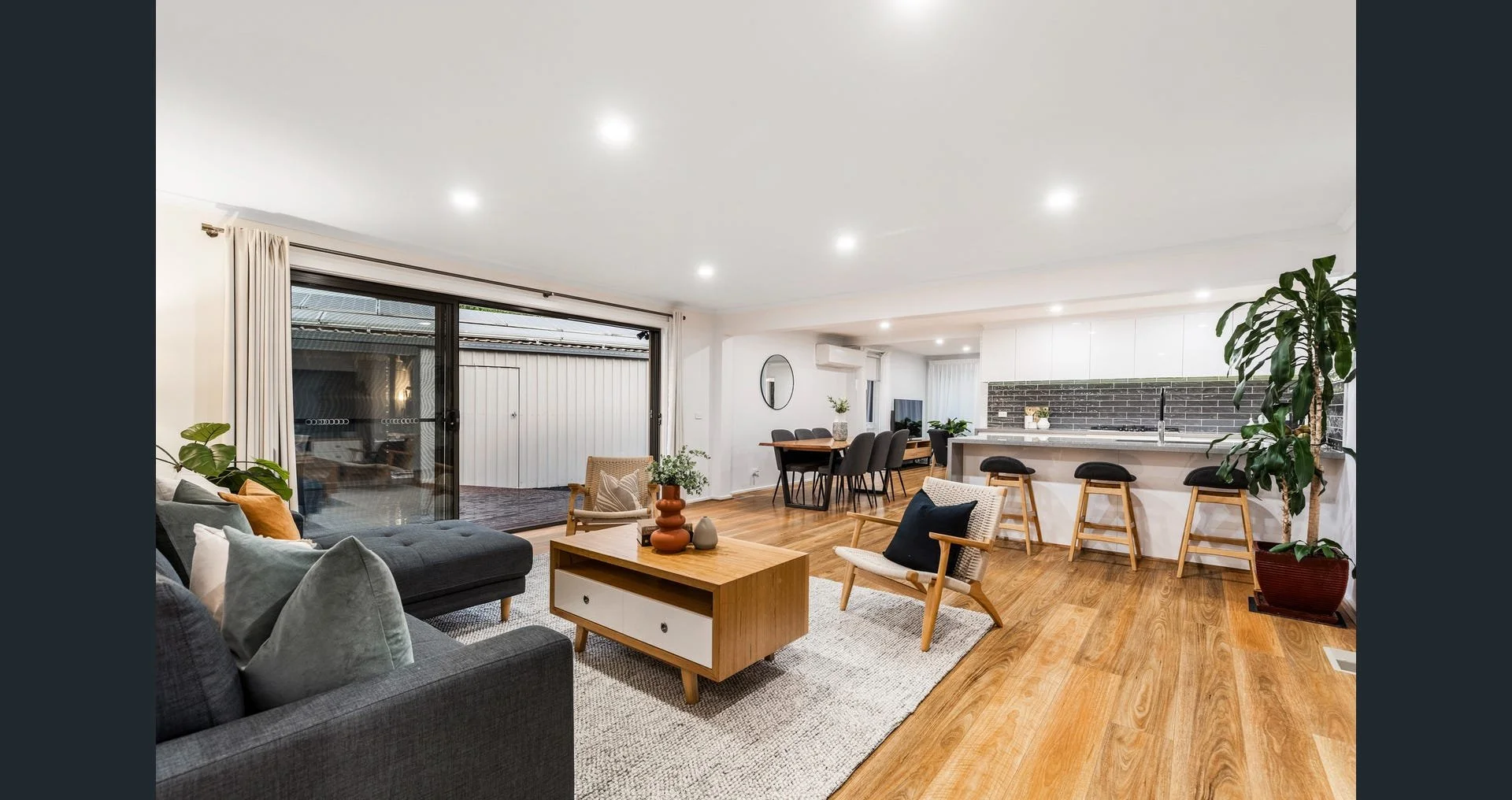 Living room with large sliding glass doors opening to outdoor patio, gray sectional sofa, wooden coffee table, armchair, and potted plants. Open-concept kitchen with white cabinets, black backsplash, bar stools, and dining area with black chairs and 