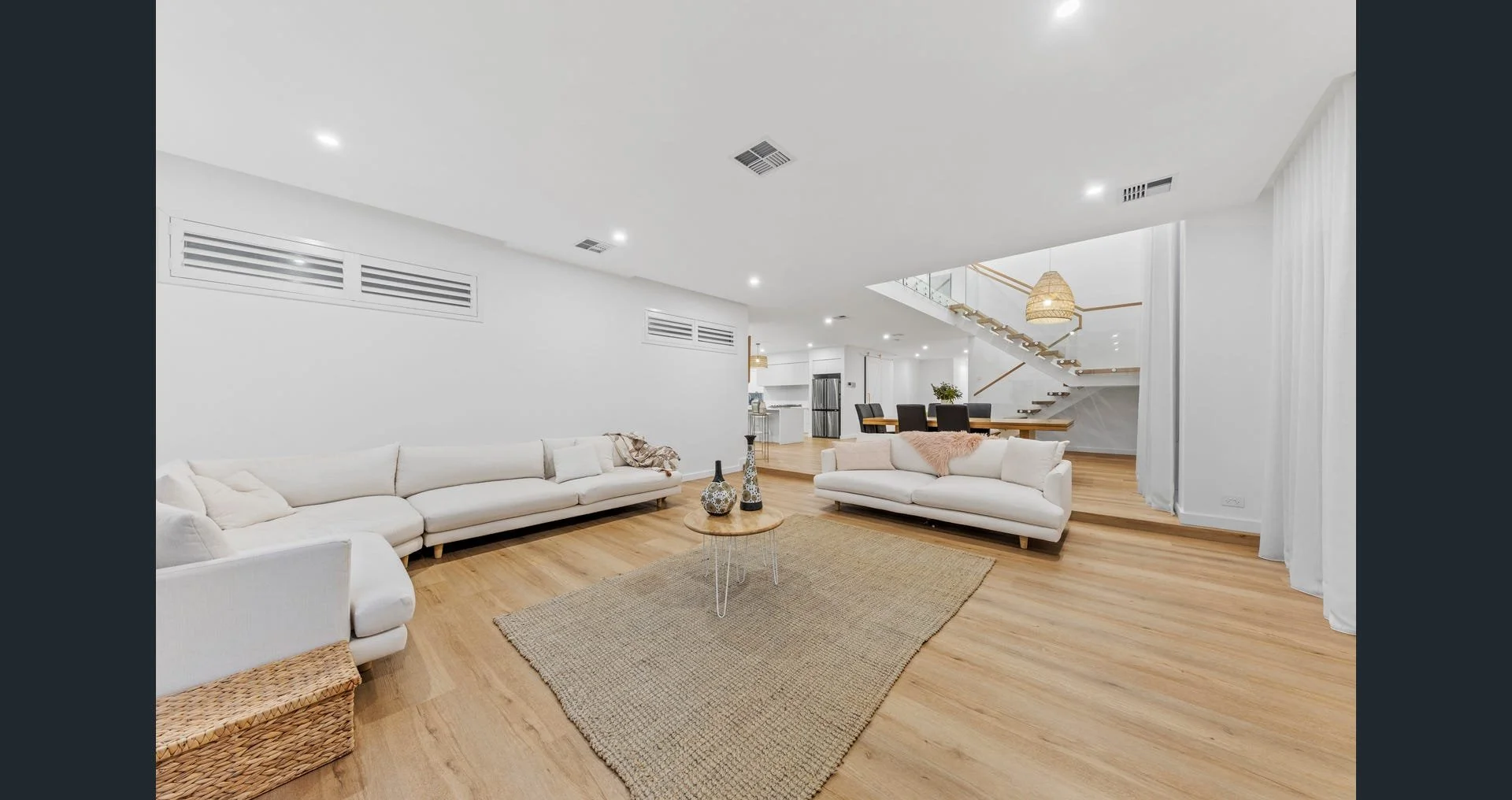 A spacious, modern living room with white sofas, decorative vases, a woven rug, hardwood floors, and a staircase in the background leading upstairs, illuminated by ceiling lights.