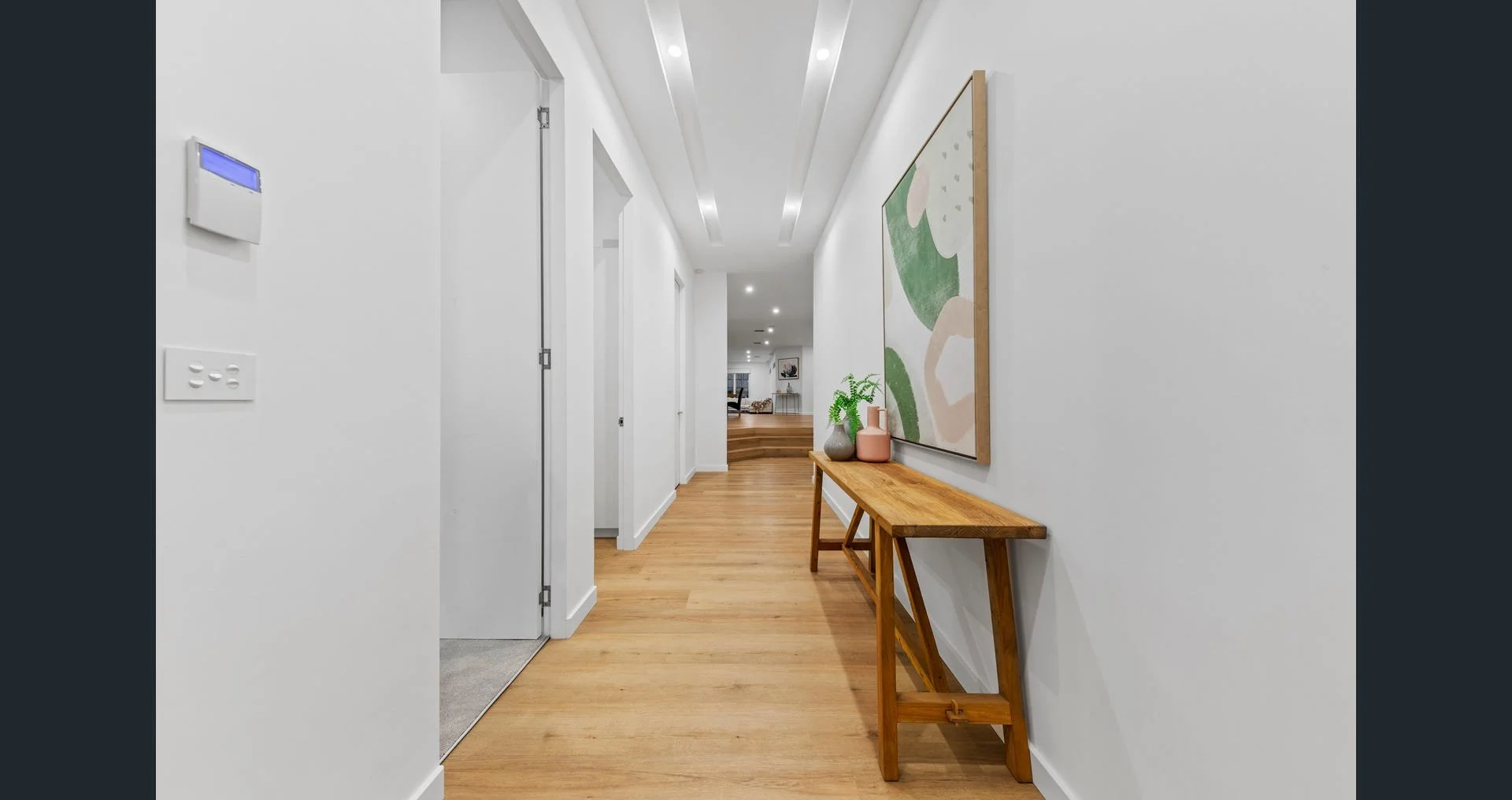 Modern hallway with white walls, wooden flooring, and recessed ceiling lights. Decor includes a wooden bench with vases and a large abstract painting on the wall.