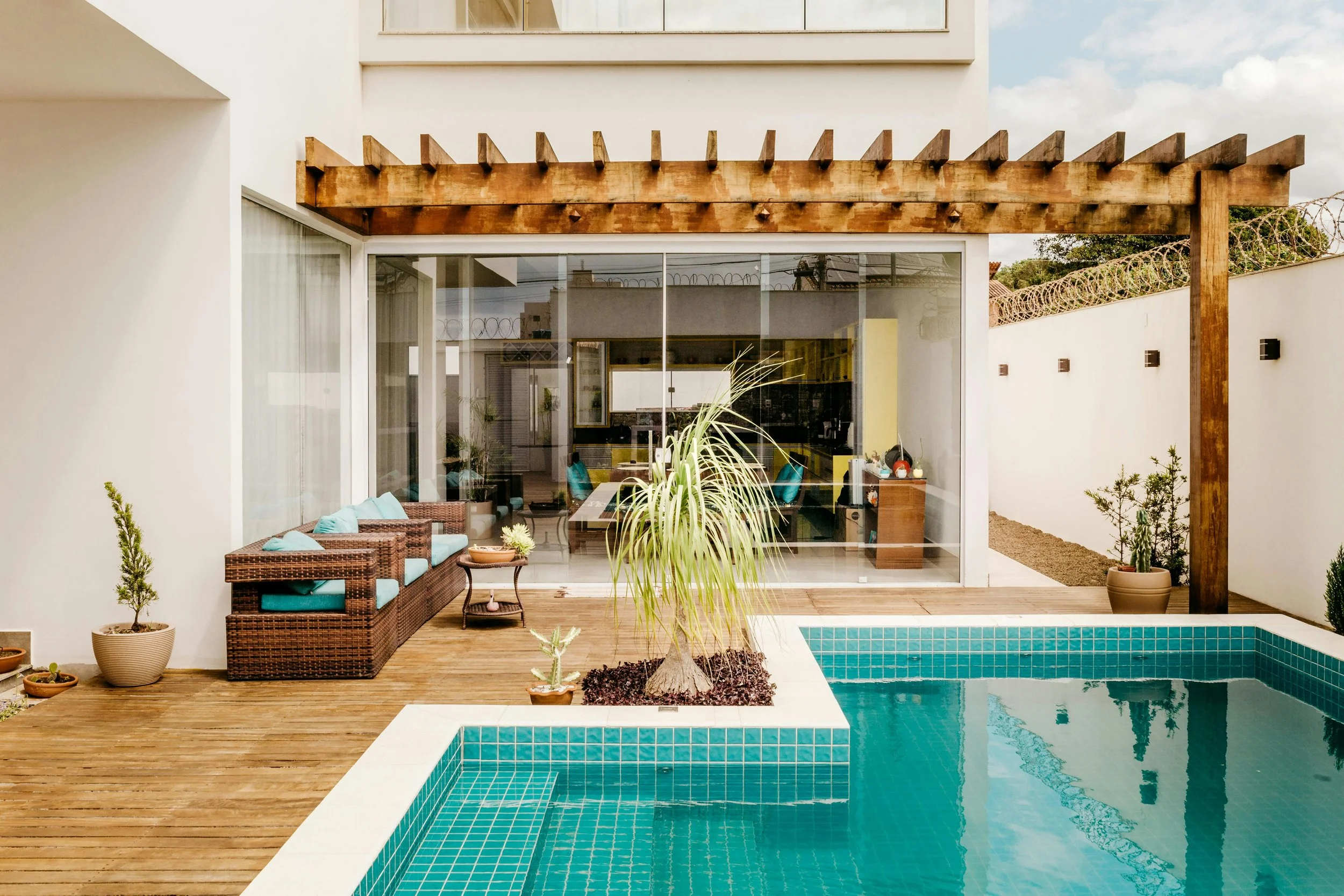 Modern backyard with a swimming pool, wooden deck, outdoor seating, potted plants, and a house with large glass sliding doors.