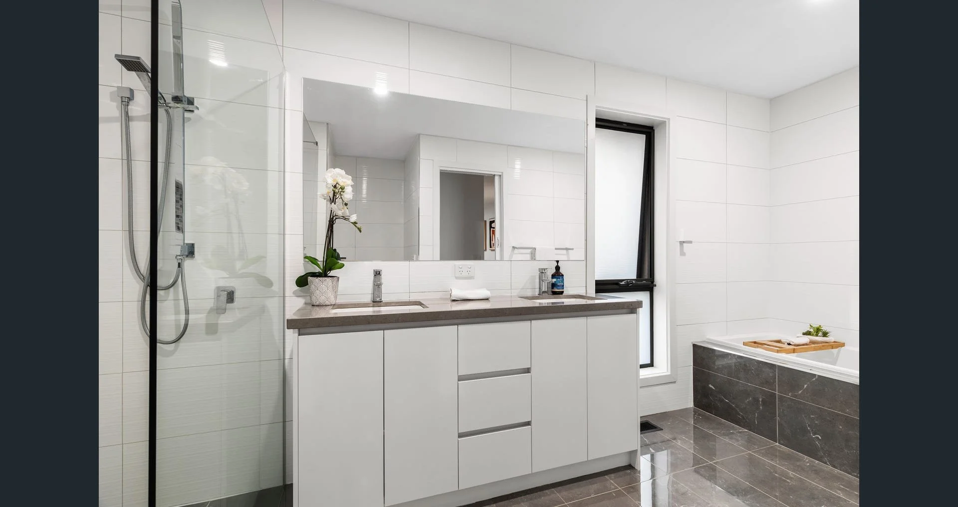 Modern bathroom with a large mirror, white cabinets, and a double sink. Features a shower area on the left with a glass partition, and a bathtub on the right with a wooden tray. The walls are tiled white and the floor has dark gray tiles.