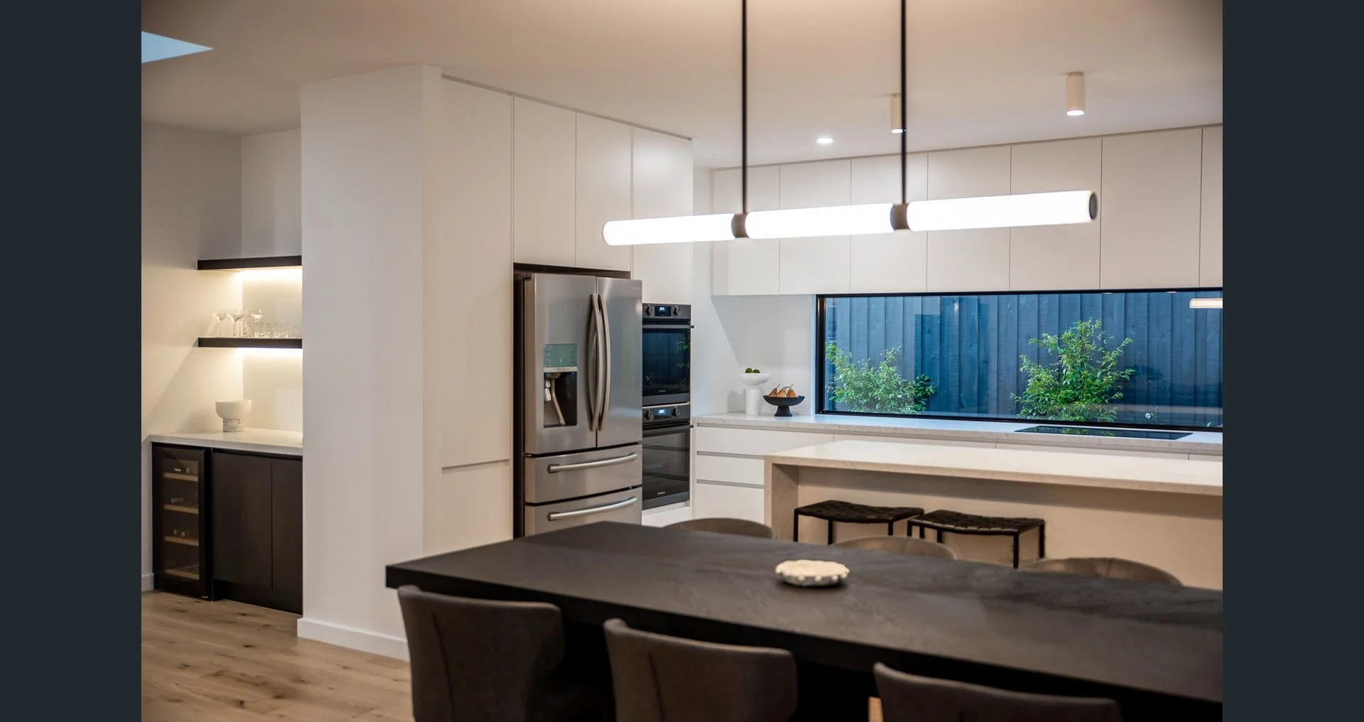 Modern kitchen with white cabinets, stainless steel refrigerator, black bar stools around a dark wood dining table, large window with outdoor greenery, and contemporary hanging light fixture.