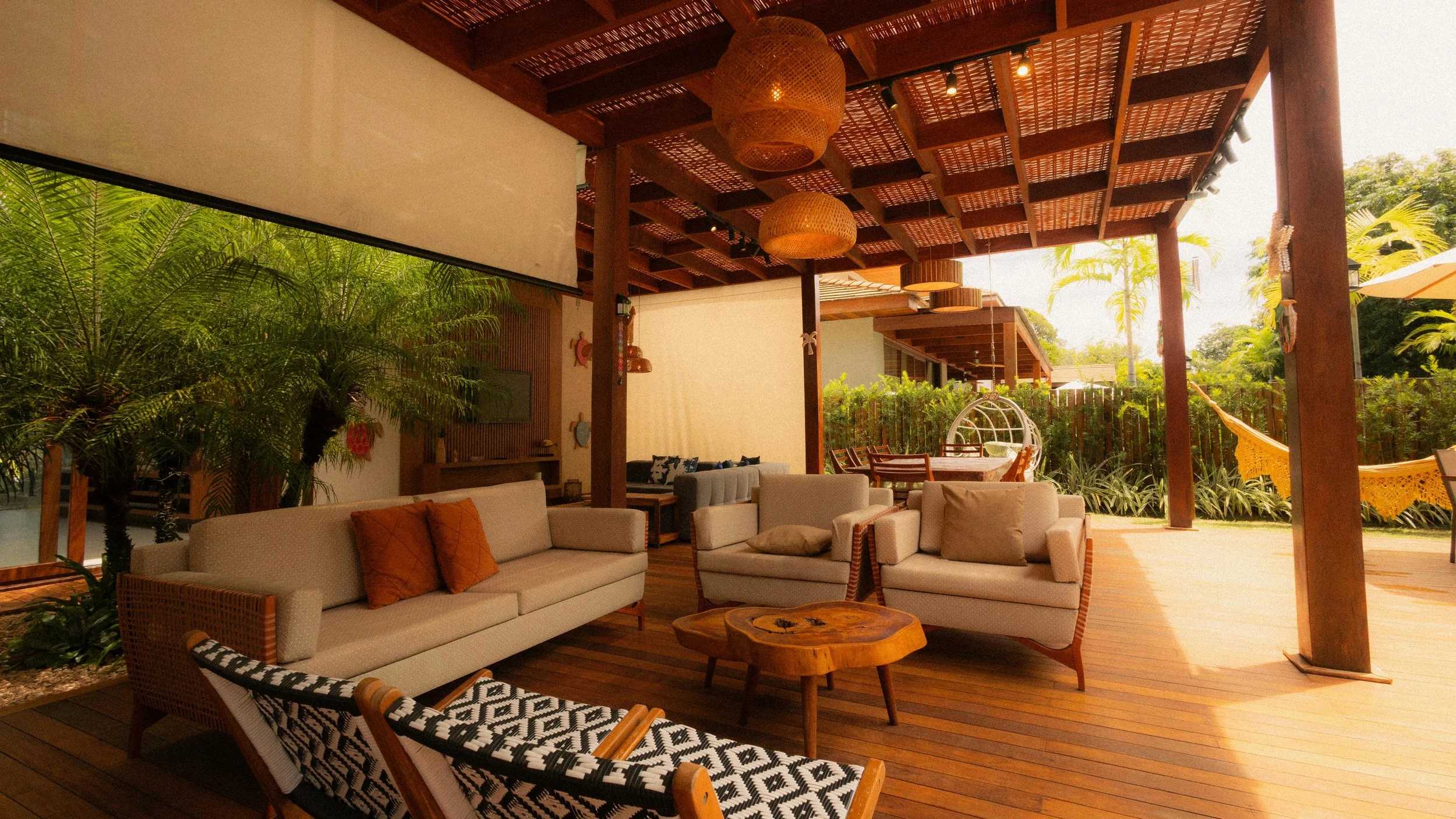 Outdoor patio with beige sofas, wooden coffee table, hanging wicker light fixtures, lush green plants, and a hammock, under a wooden pergola with roofing, sunlight filtering through, surrounded by palm trees.