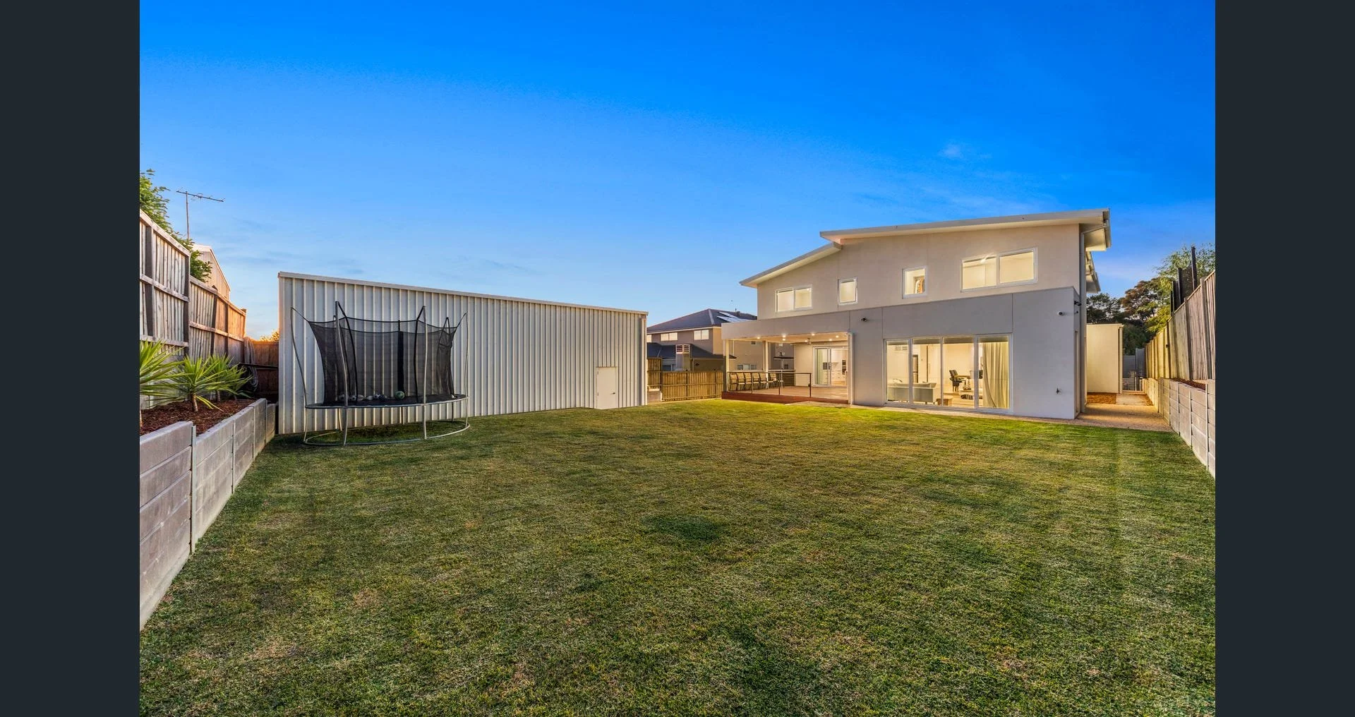 Backyard with lawn, a trampoline on the left, a modern two-story house on the right, enclosed by a wooden fence, under a clear blue sky at sunset.