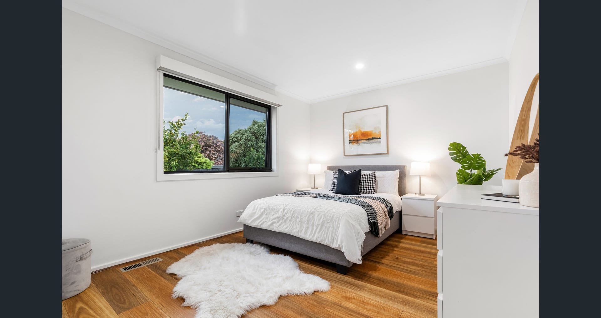 A modern bedroom with white walls, a large window showing trees outside, a bed with white bedding and black pillows, two nightstands with lamps, a framed artwork, a white dresser with plants and decor, a fluffy white rug, and hardwood flooring.