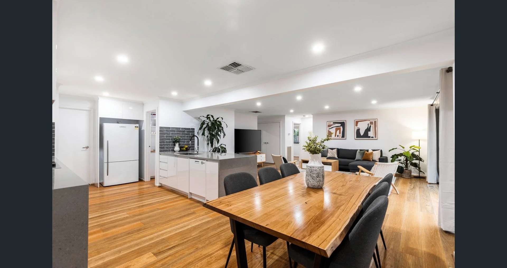 Open-concept living space with a kitchen and dining area, featuring white cabinetry, a wooden dining table with black chairs, a black sofa, wall art, and potted plants, illuminated by recessed ceiling lights.