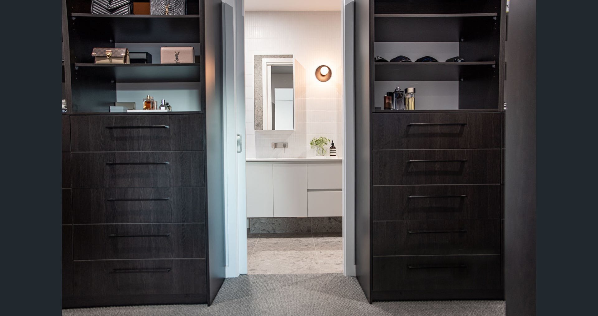 A doorway with dark wooden sliding closet doors open to reveal a bathroom with white vanity, mirror, and decorative items, viewed from a room with carpeted flooring.