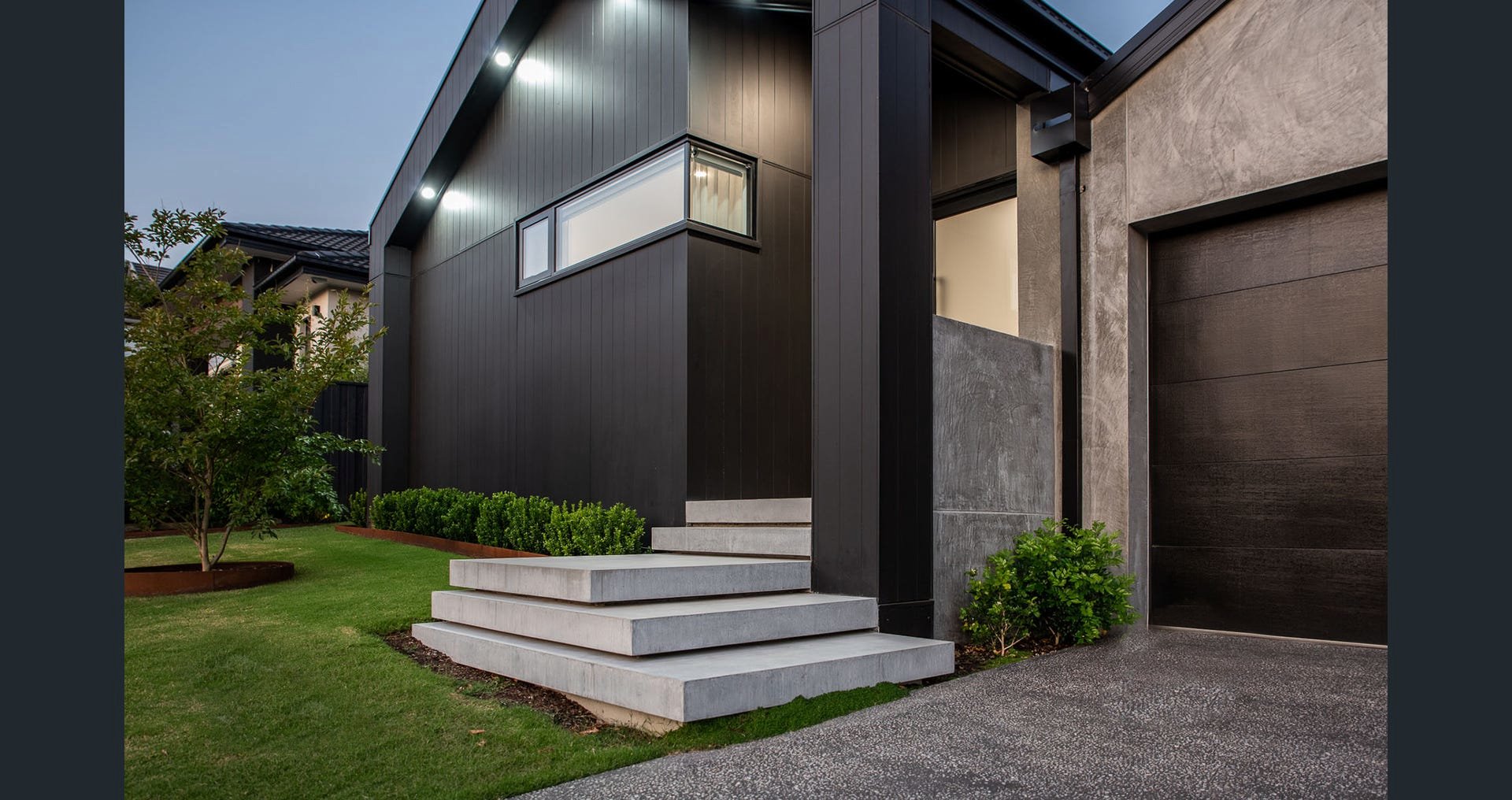 Modern house exterior with black siding, concrete steps, a brown garage door, green shrubs, and manicured lawn during dusk.