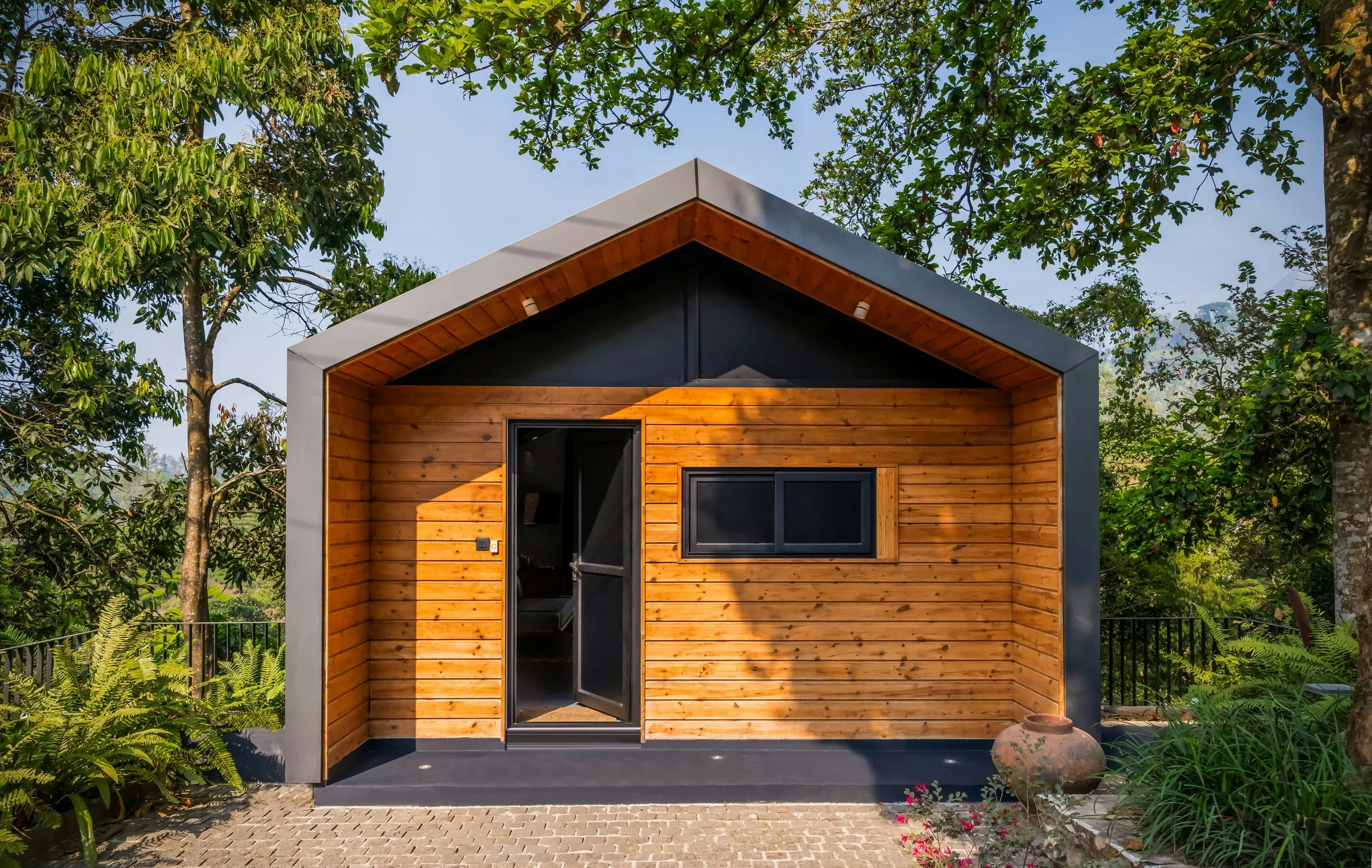 A modern tiny house with a wood exterior and black trim, surrounded by lush green trees and plants.