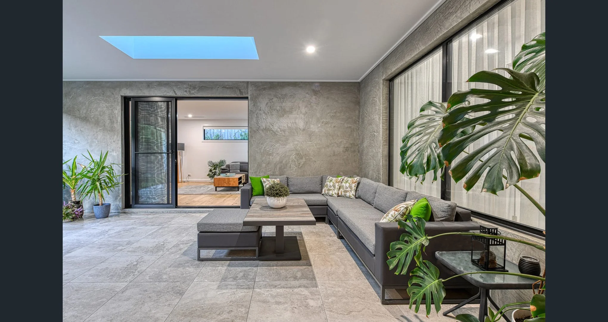 Modern living room with gray L-shaped sofa, green cushions, and large potted plants, with natural light from a skylight and windows.