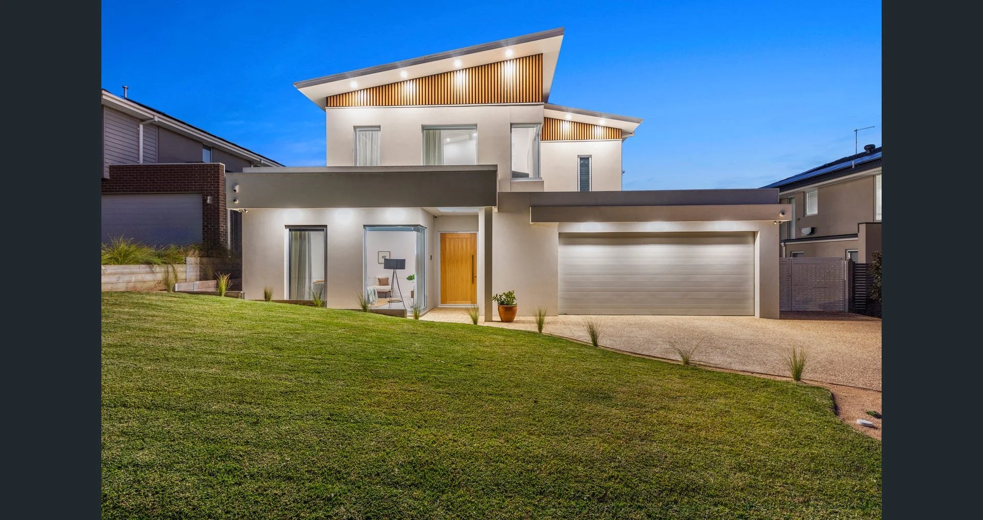 Modern two-story house with a landscaped lawn, illuminated windows, and a closed garage door during dusk.
