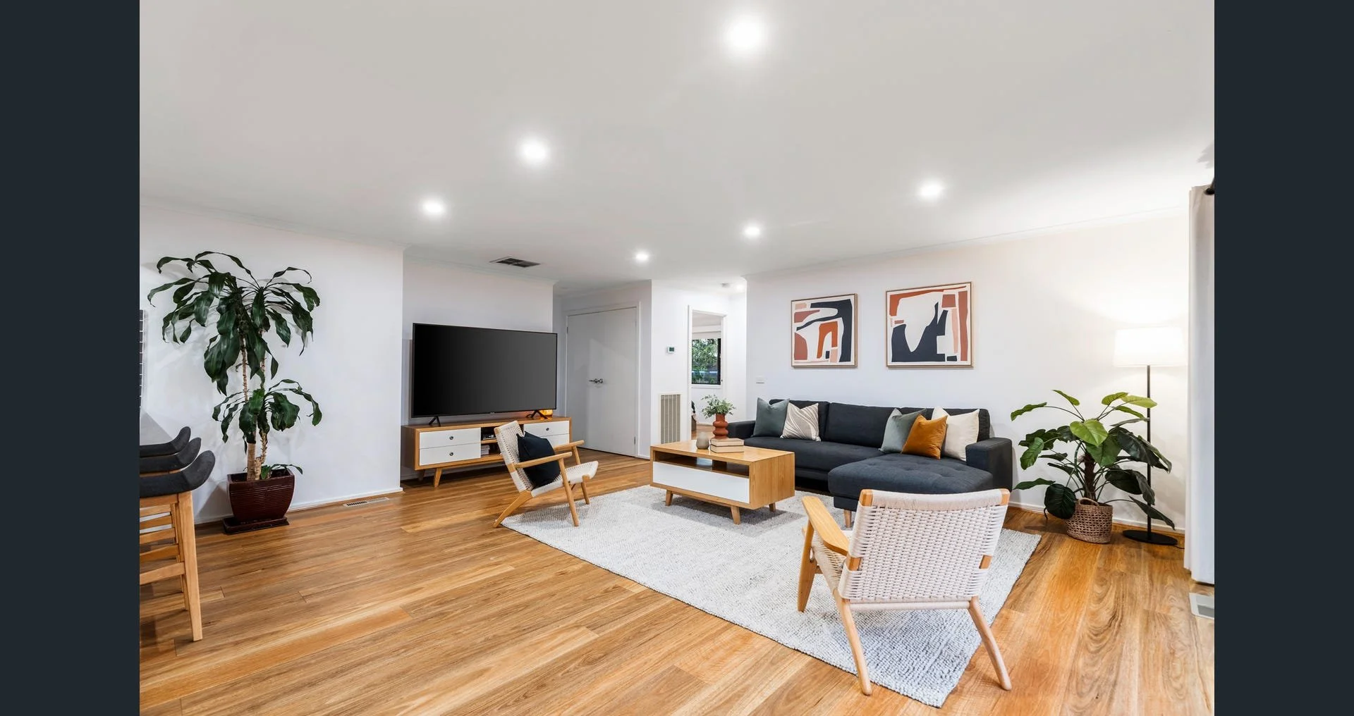 A modern living room with hardwood floors, white walls, and recessed ceiling lights. It features a black sofa with multiple pillows, a light-colored armchair, and a wooden coffee table. There are two abstract art pieces hanging on the wall, a large p