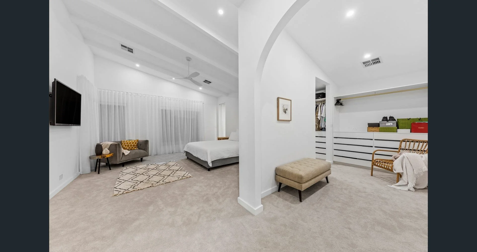 A spacious modern bedroom with white walls and ceiling, beige carpet, and minimal decor. The room is divided into a sleeping area with a bed, and a sitting or dressing area with a chair, bench, and storage. Windows with sheer white curtains are on th