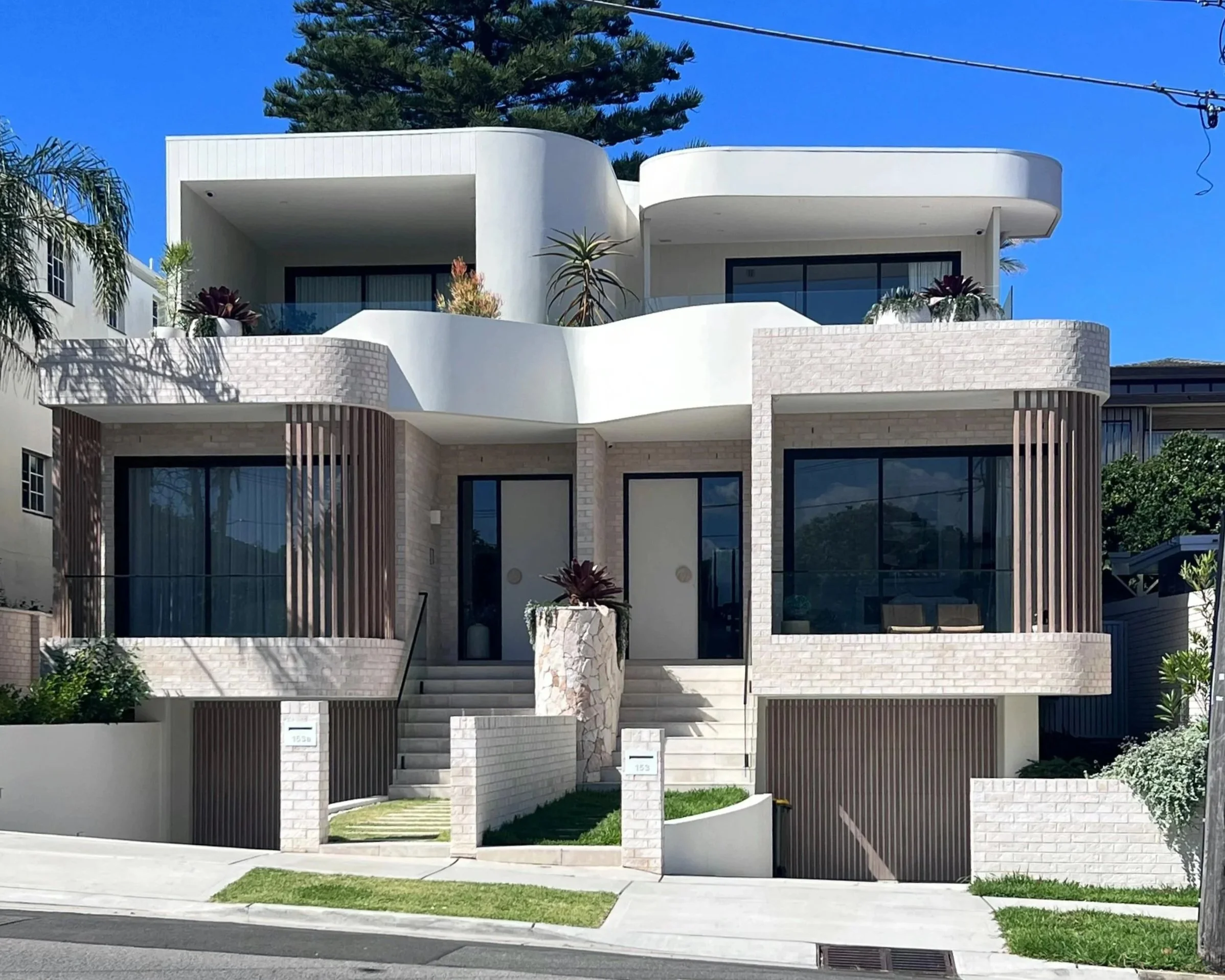 A modern white multi-story house with curved architectural design, large glass windows, and a small front yard with stairs leading to the entrance. There are plants and trees in the background and a clear blue sky.