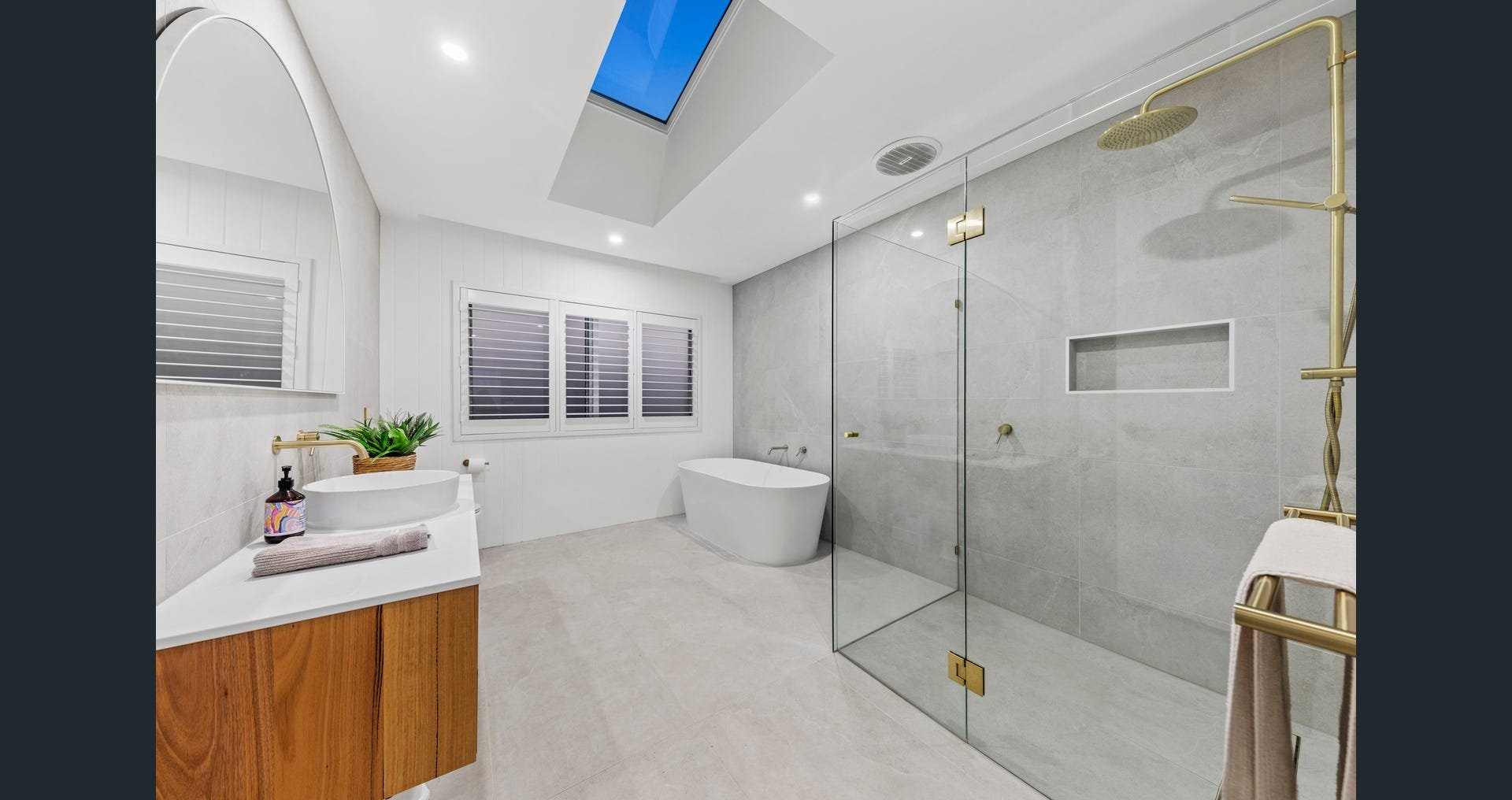 Modern bathroom with white walls, a bathtub, a glass shower enclosure with gold fixtures, a wooden vanity with a vessel sink, a large window with blinds, and a skylight.