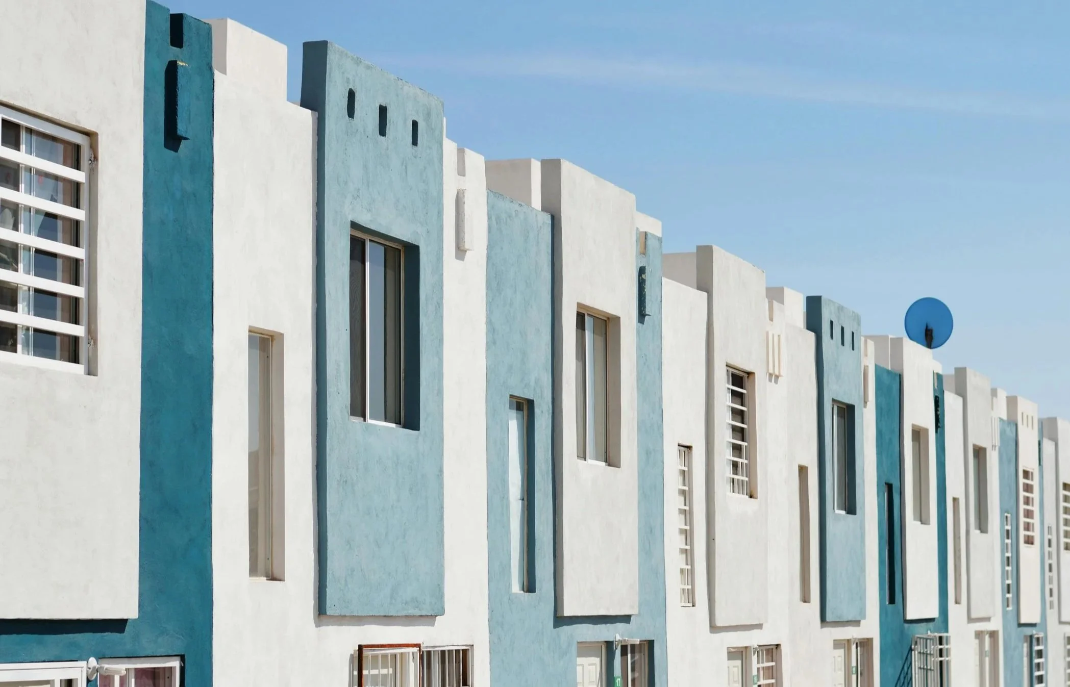 Modern apartment building with white and light blue exterior, multiple rectangular windows, and a clear blue sky in the background.