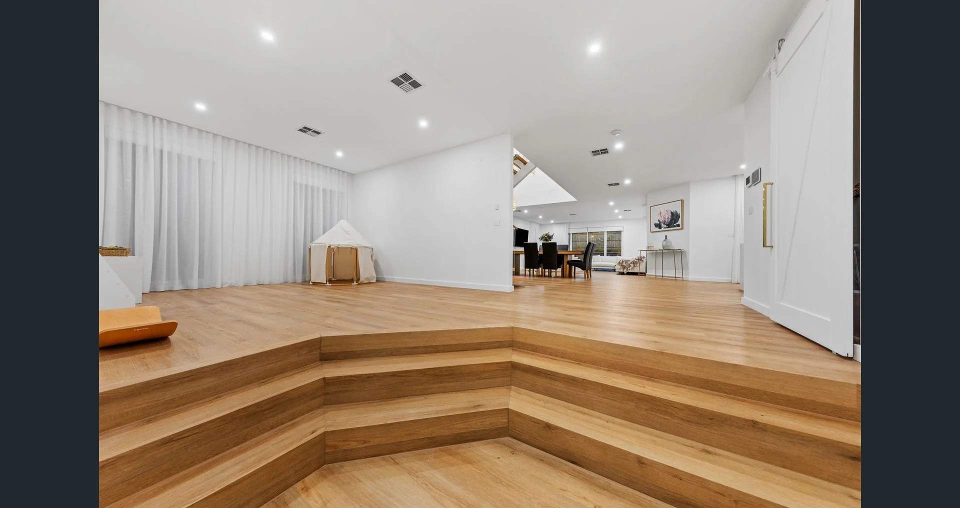Modern open-plan living space with wooden flooring, white walls, and recessed lighting, featuring a small raised platform with curved steps.