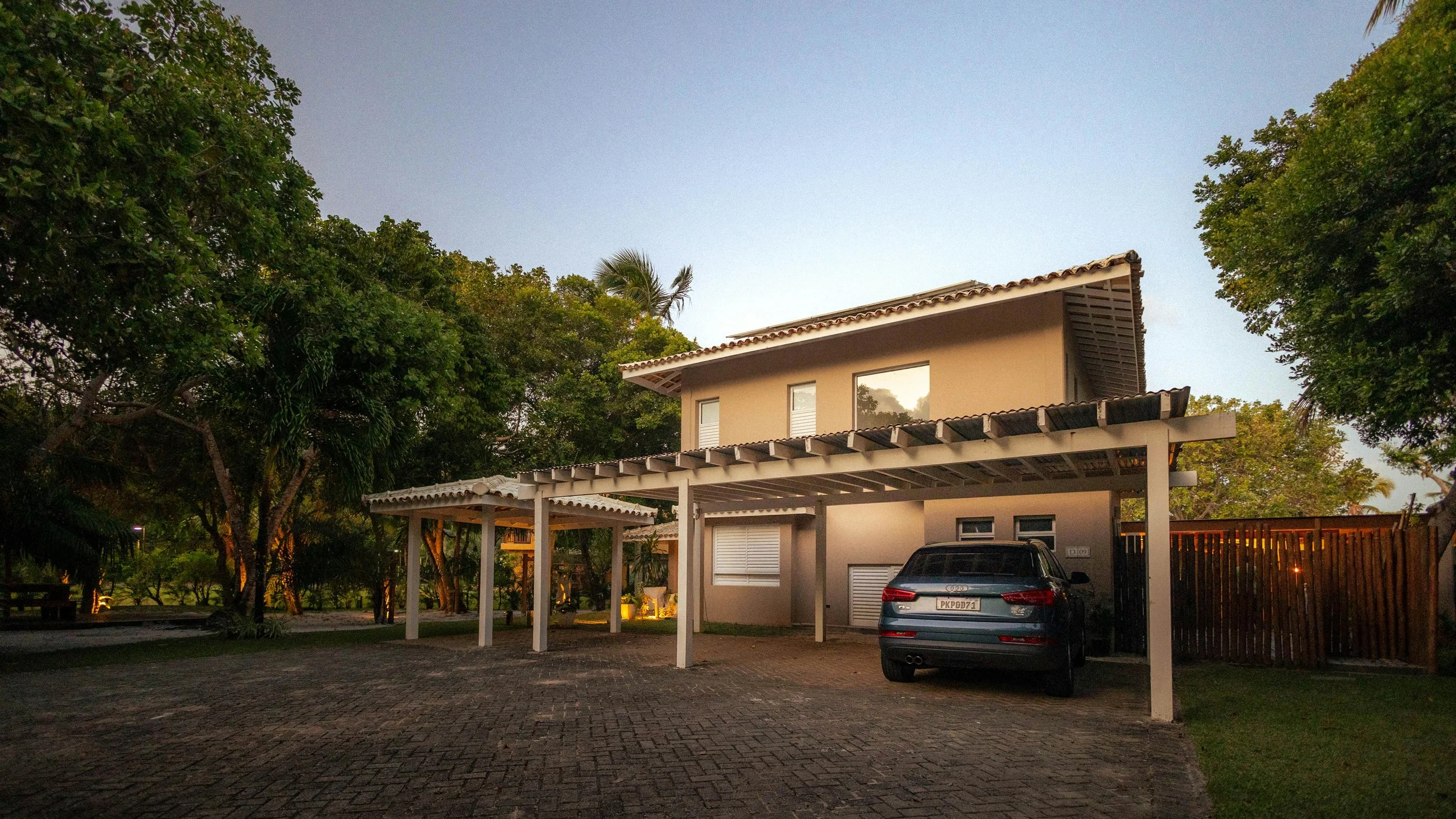 A modern two-story house with a car parked under a white carport, surrounded by trees and a wooden fence, during dusk.