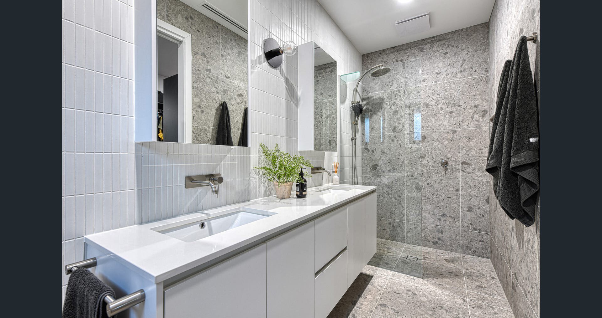 Modern bathroom with a double sink vanity, large mirrors, gray marble tiles in the shower area, and black towels hanging on the wall.