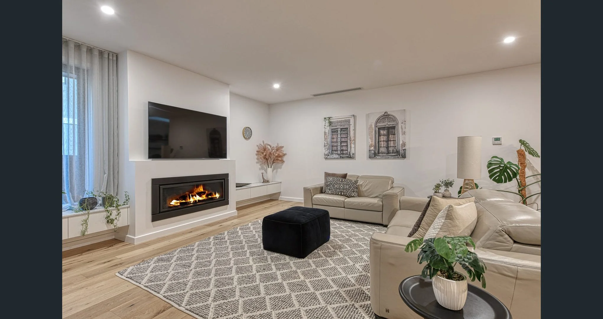 Living room with beige sofas, wood flooring, wall-mounted TV, fireplace, artwork, plants, and a patterned rug.