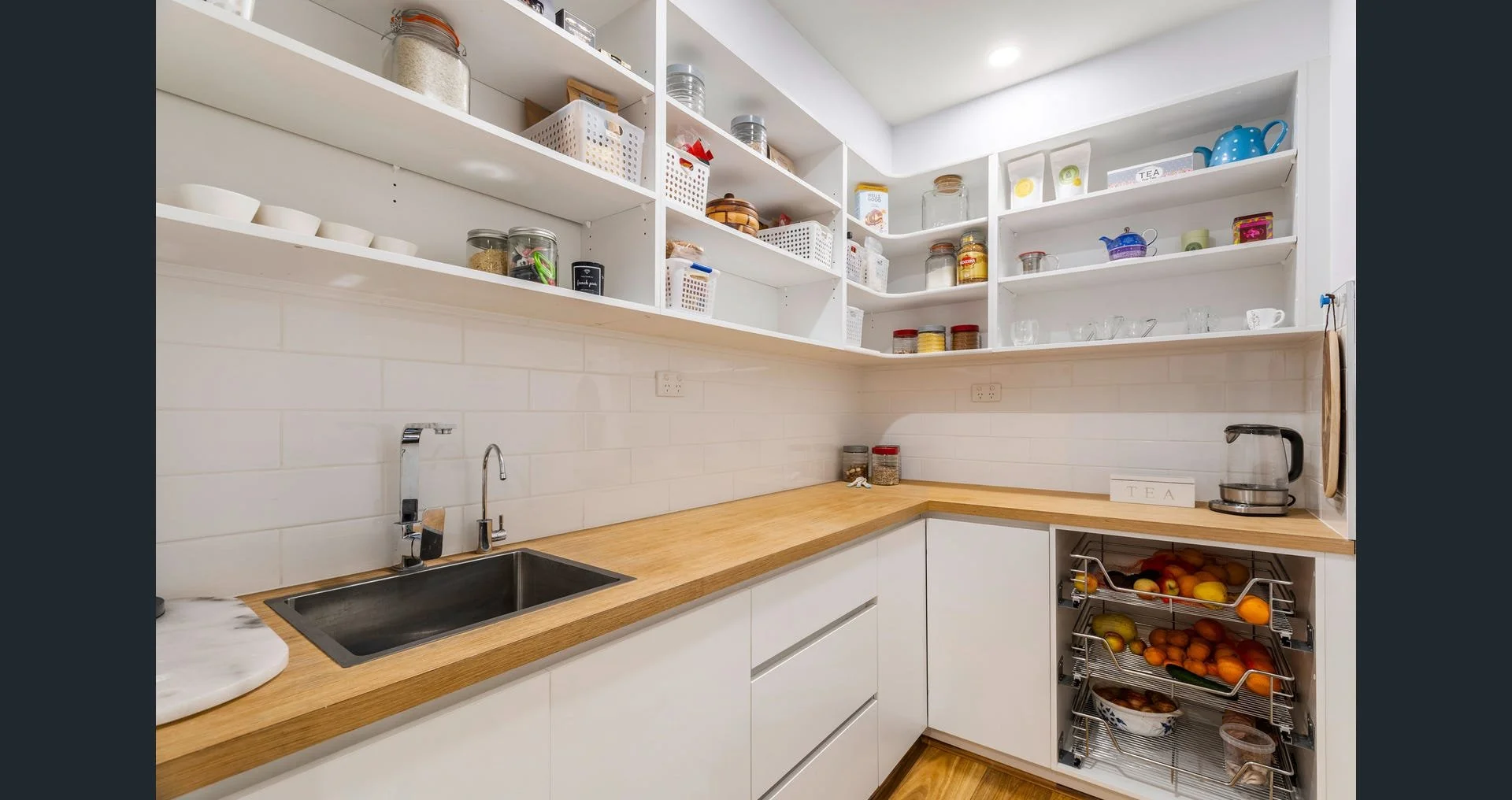 A modern kitchen with white cabinets, open shelves filled with jars, cups, and containers, a wooden countertop, a sink, a kettle, and a two-tiered fruit basket with oranges and apples.