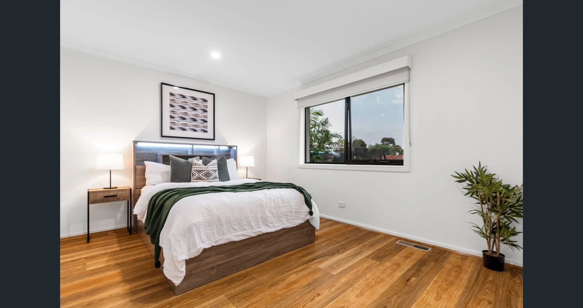 A bedroom with a wooden bed frame, white bedding, and green throw blanket. There are nightstands with lamps on each side of the bed, and a framed abstract art piece above the headboard. The room has a large window with a view of trees outside, a pott